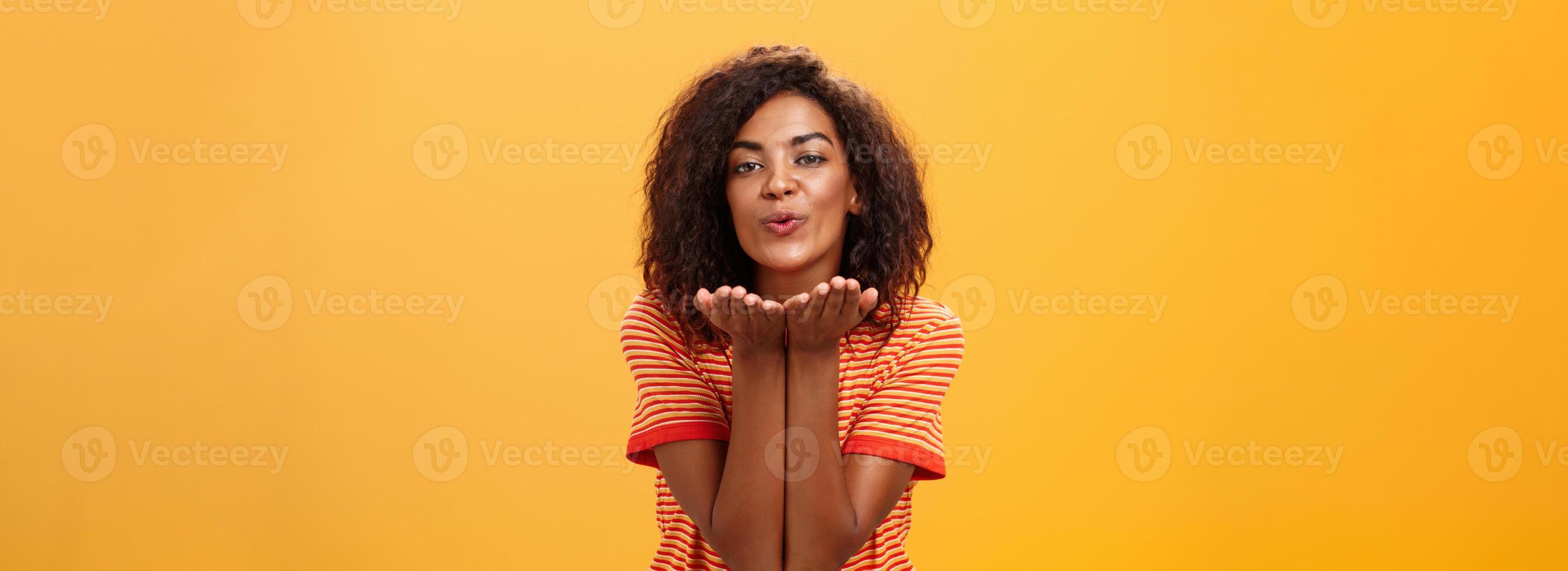 Sending passionate kiss to most loving person. Romantic attractive and stylish young african american girlfriend with curly hairstyle bending towards camera with slight smile, folded lips blowing mwah photo