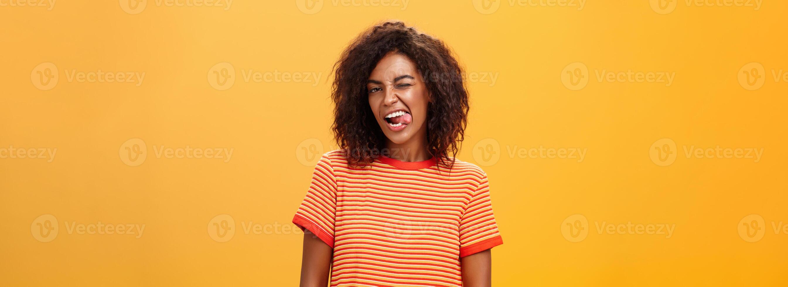 Portrait of daring and emotive confident flirty woman with afro hairstyle winking joyfully showing tongue posing carefree and enthusiastic against orange background flirting with hot guy photo