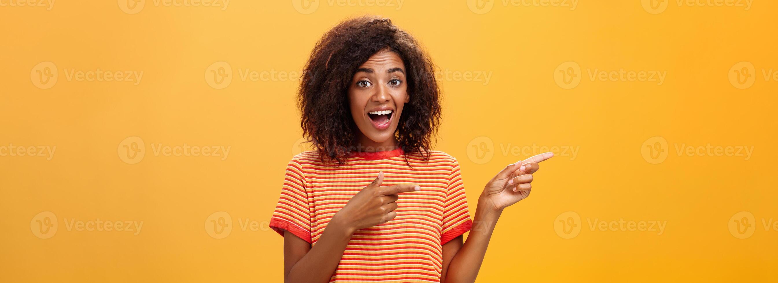 Portrait of amazed excited charismatic dark-skinned young pretty girl with afro hairstyle in trendy striped t-shirt pointing left delighted and fascinated posing against orange background photo