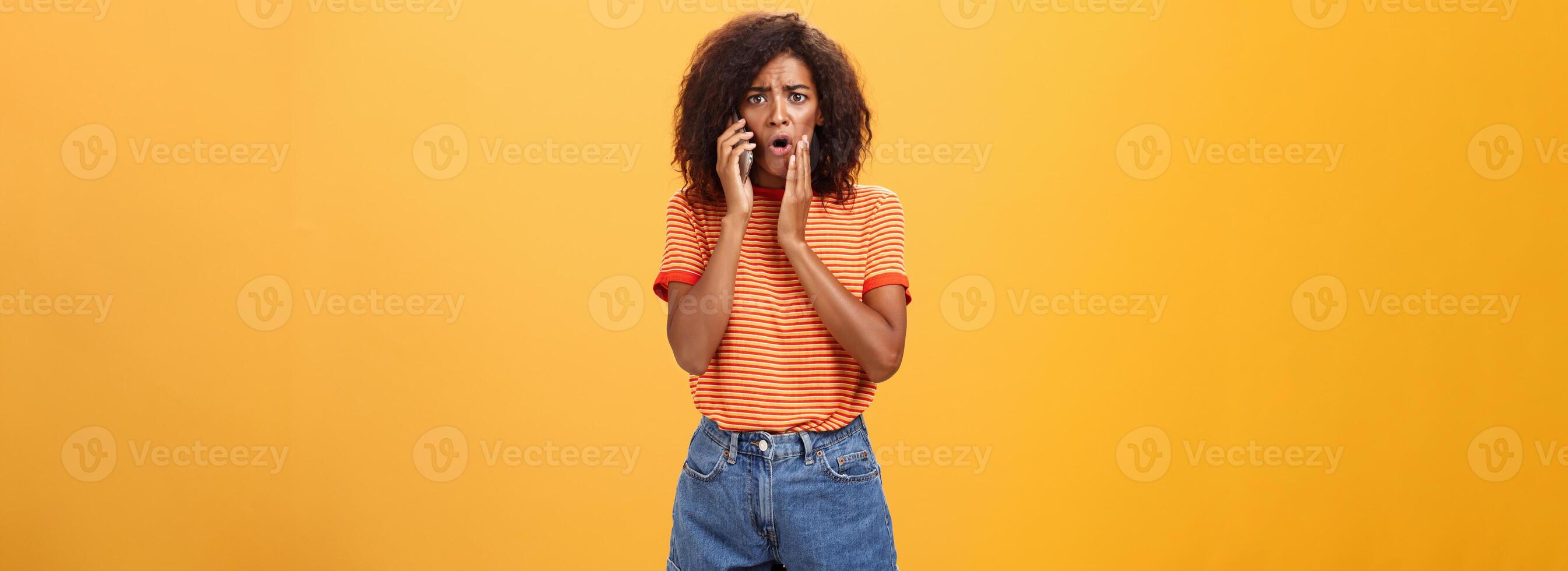 Woman receiving disturbing call feeling emapthy and sorry for poor friend getting in trouble holding cellphone near ear touching cheek concerned frowning standing sad, worried over orange background photo