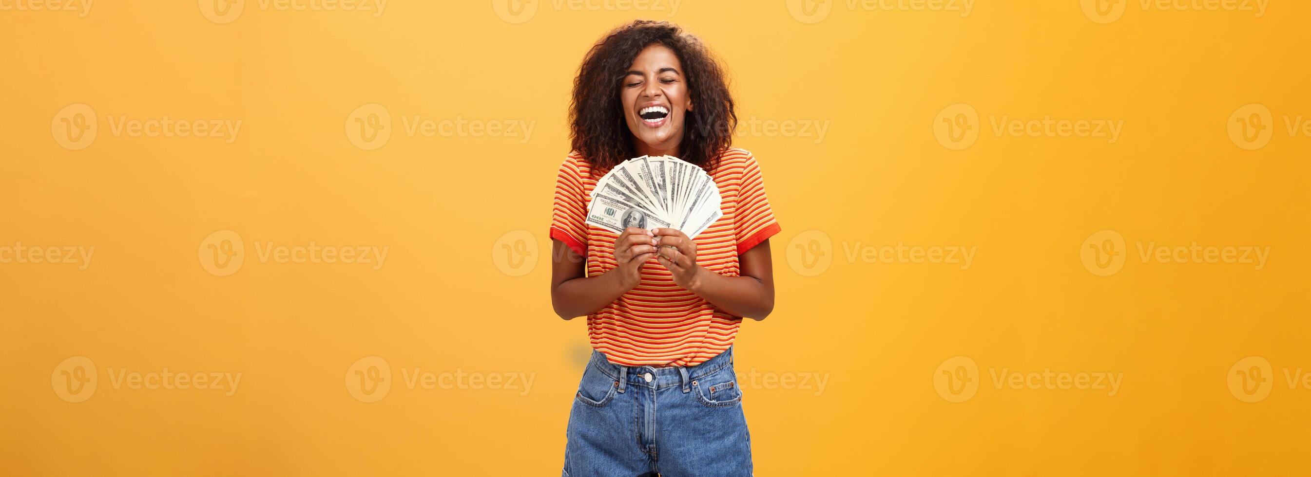 finalmente niña rico. retrato de contento carismático elegante joven hembra modelo recepción primero pago participación un montón de dinero y riendo desde alegría victorioso lotería Listo residuos efectivo terminado naranja antecedentes foto