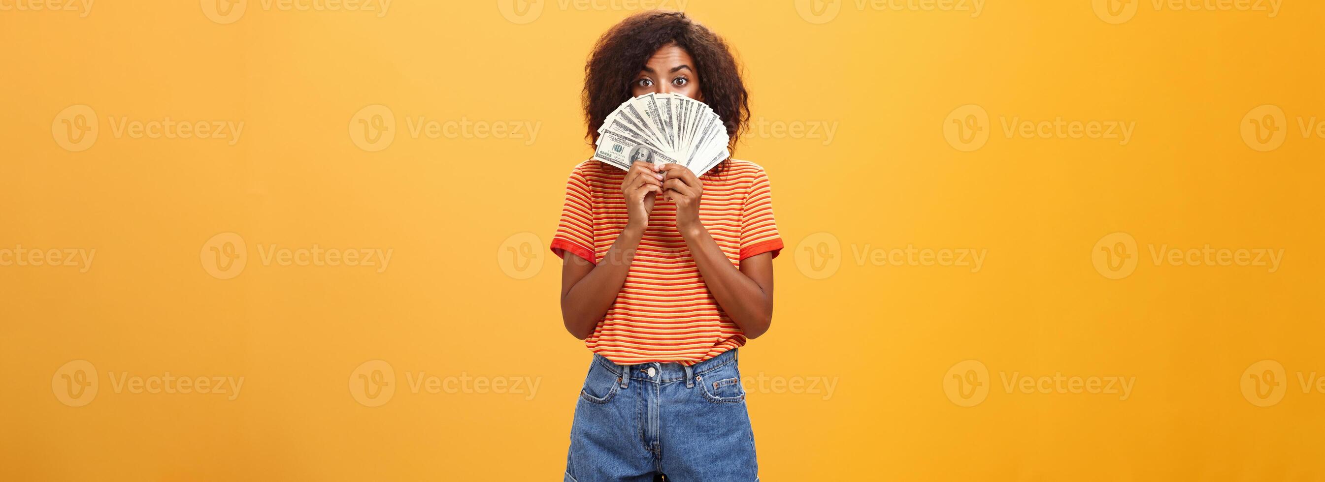 Woman gonna waste lots of cash today. Pleased and delighted rich african american girl with curly hairstyle hiding face behind bunch of money peeking mysteriously at camera winning millions photo