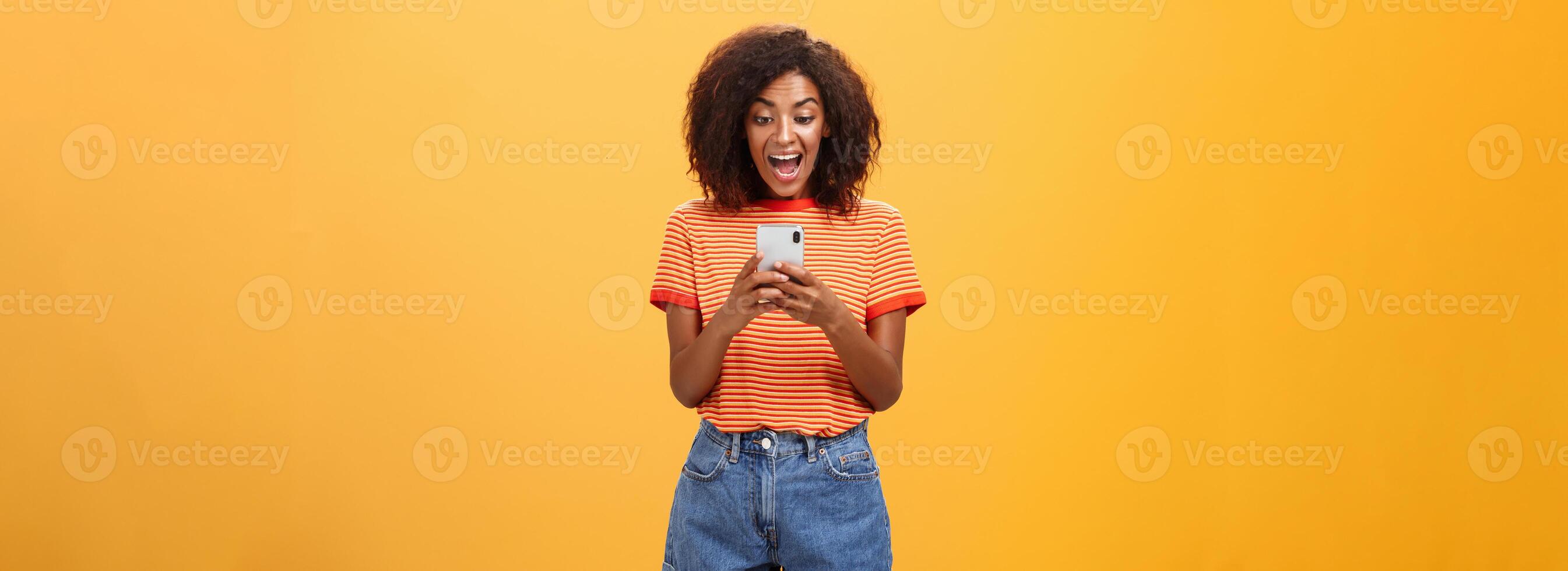 Girl expressing excitement and joy receiving awesome invitation via messages yelling from delight and happiness looking at smartphone screen impressed and thrilled over orange background photo