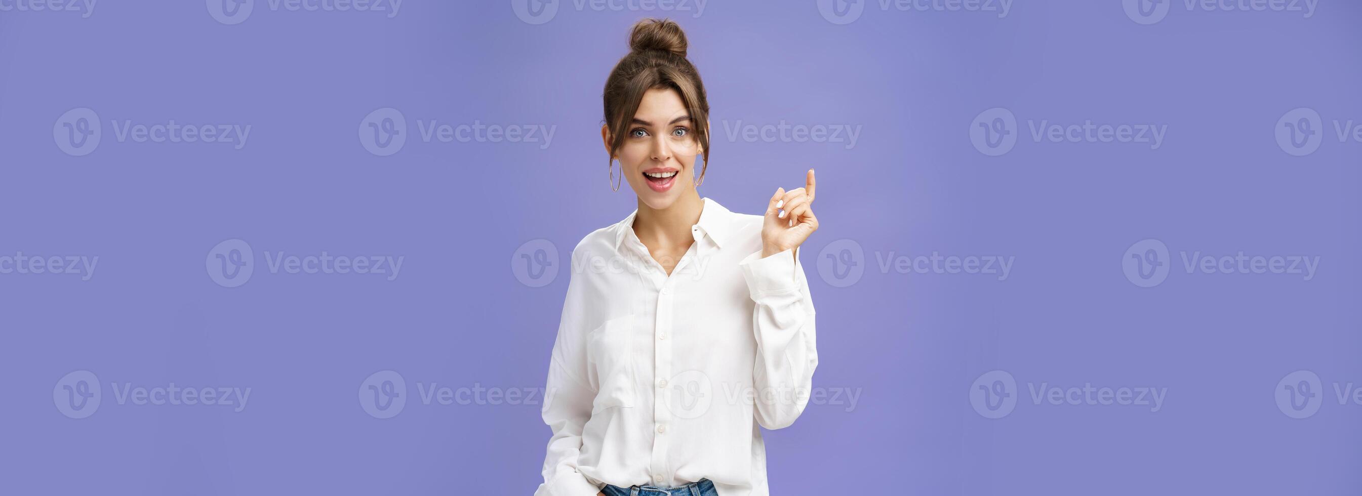 Expressive charismatic and beautiful white girl with combed hair in stylish white blouse holding hand in pocket gesturing during talk and smiling at camera with friendly gaze over purple wall photo