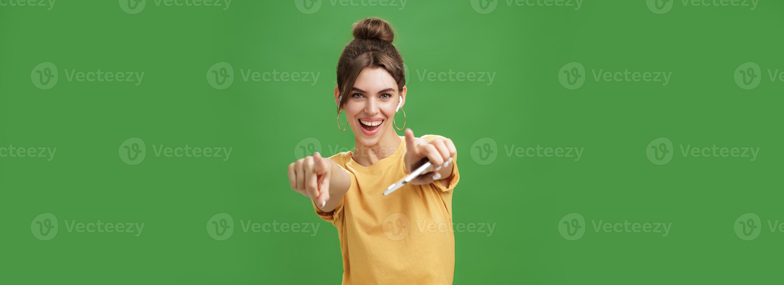 energizado carismático atractivo joven mujer con cardado pelo y linda diastema participación teléfono inteligente bailando alegremente escuchando música en inalámbrico auriculares señalando y sonriente a cámara terminado verde pared foto