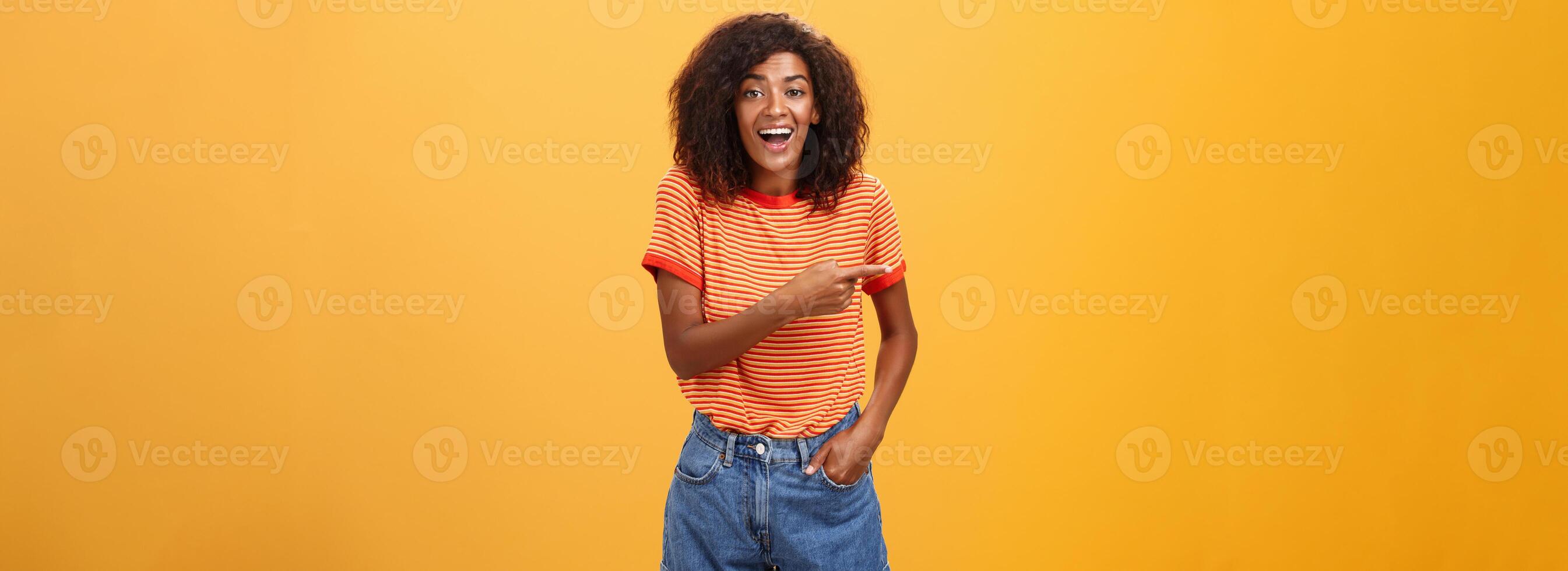 mujer riendo terminado gracioso cachorro. retrato de divertido y entretenido atractivo elegante afroamericano hembra en casual mezclilla pantalones cortos señalando izquierda hablando acerca de increíble Copiar espacio terminado naranja pared foto