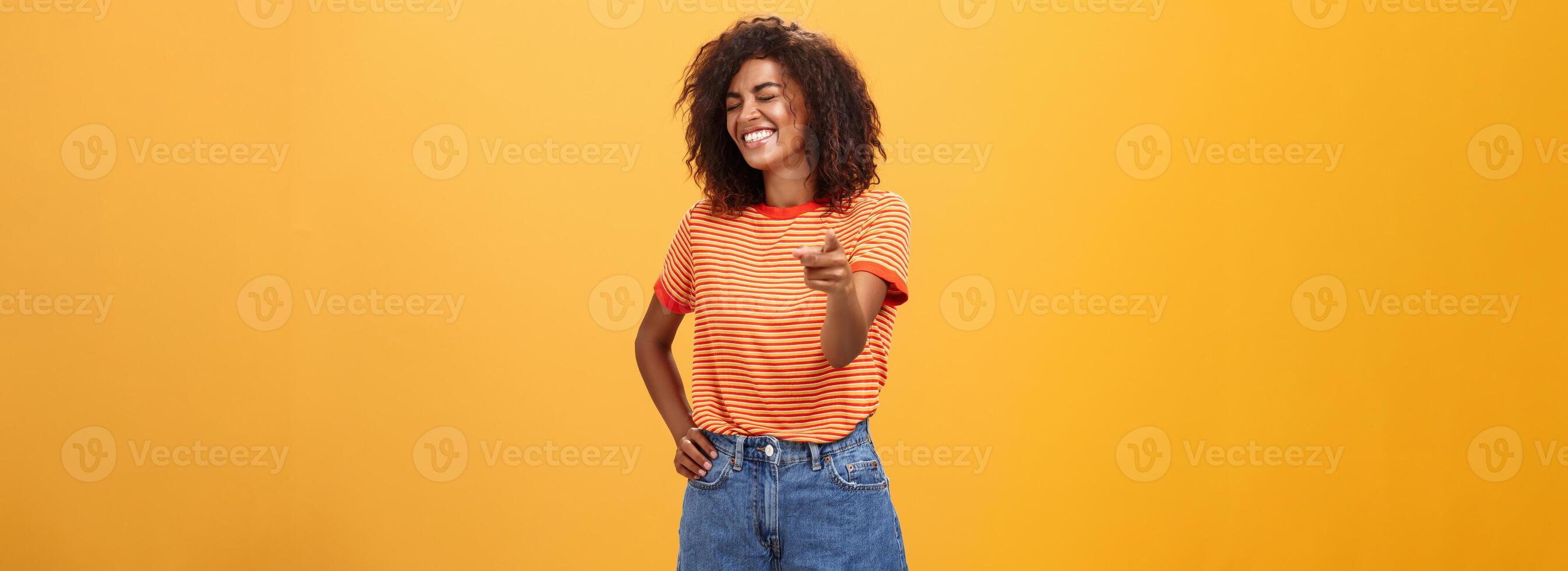 Girl pointing at funny and hilarious friend approving good joke. Amused and entertained attractive dark-skinned female with afro hairstyle laughing with closed eyes indicating at camera joyfully photo