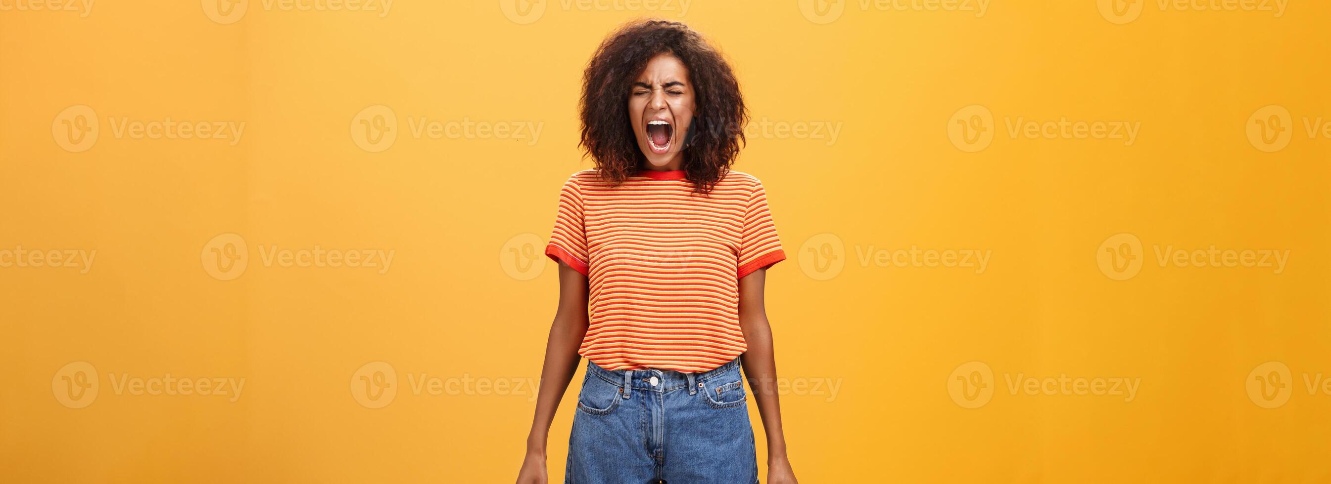 Girl feeling distressed and tensed losing temper and control of feelings yelling out loud clenching fists closing eyes standing intense and depressed over orange background screaming from rage photo