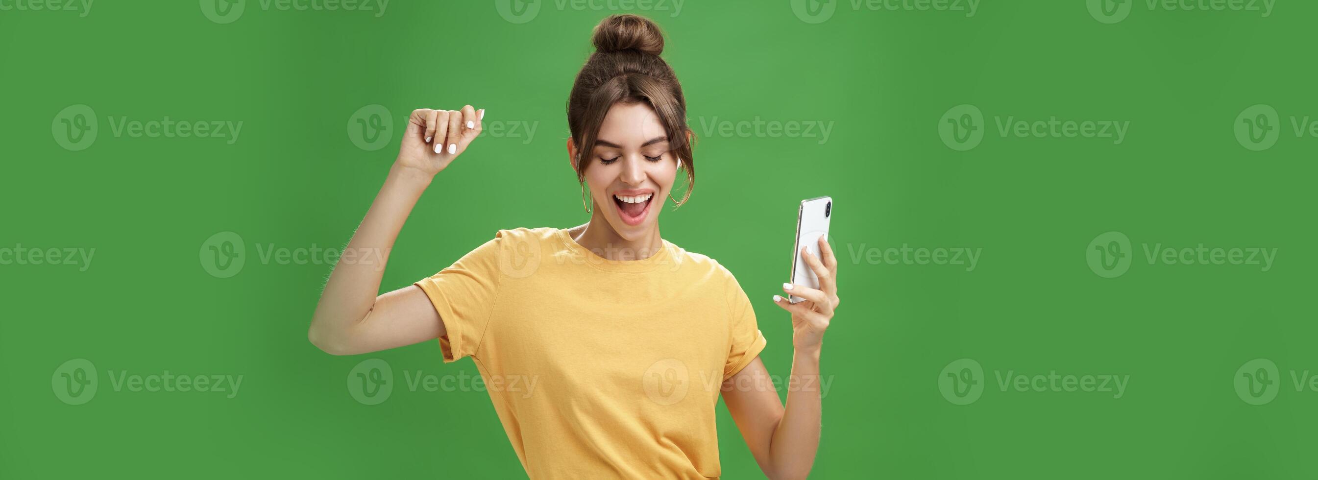 frio y elegante contento mujer con tatuaje y brecha dientes en amarillo camiseta bailando alegremente escuchando música en inalámbrico auriculares participación teléfono inteligente clausura ojos y sonriente desde deleite terminado verde pared foto