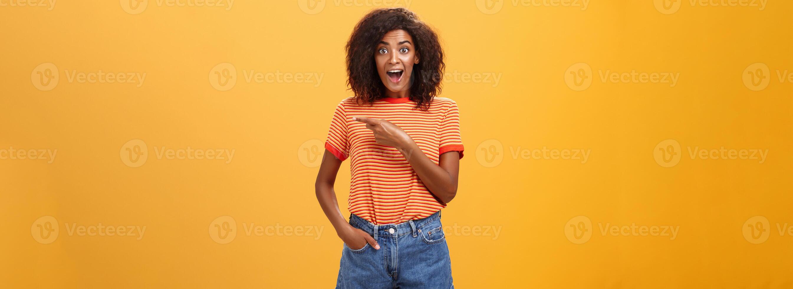 Hey awesme copy space there. Portrait of impressed and surprised enthusiastic young african american female with afro hairstyle pointing right astonished looking energized and curious over orange wall photo