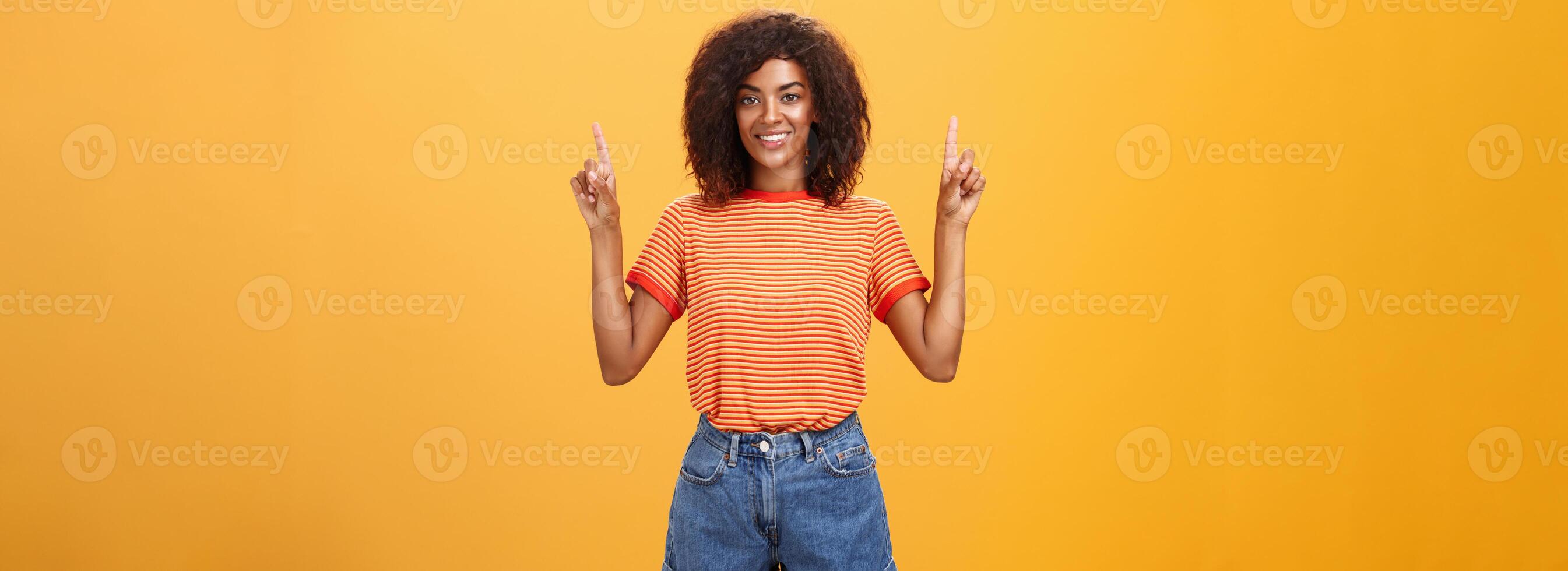 mirando solamente arriba y adelante. optimista ambicioso elegante de piel oscura hembra estudiante en a rayas frio camiseta y pantalones cortos levantamiento manos señalando hacia arriba y sonriente simpático terminado naranja pared foto