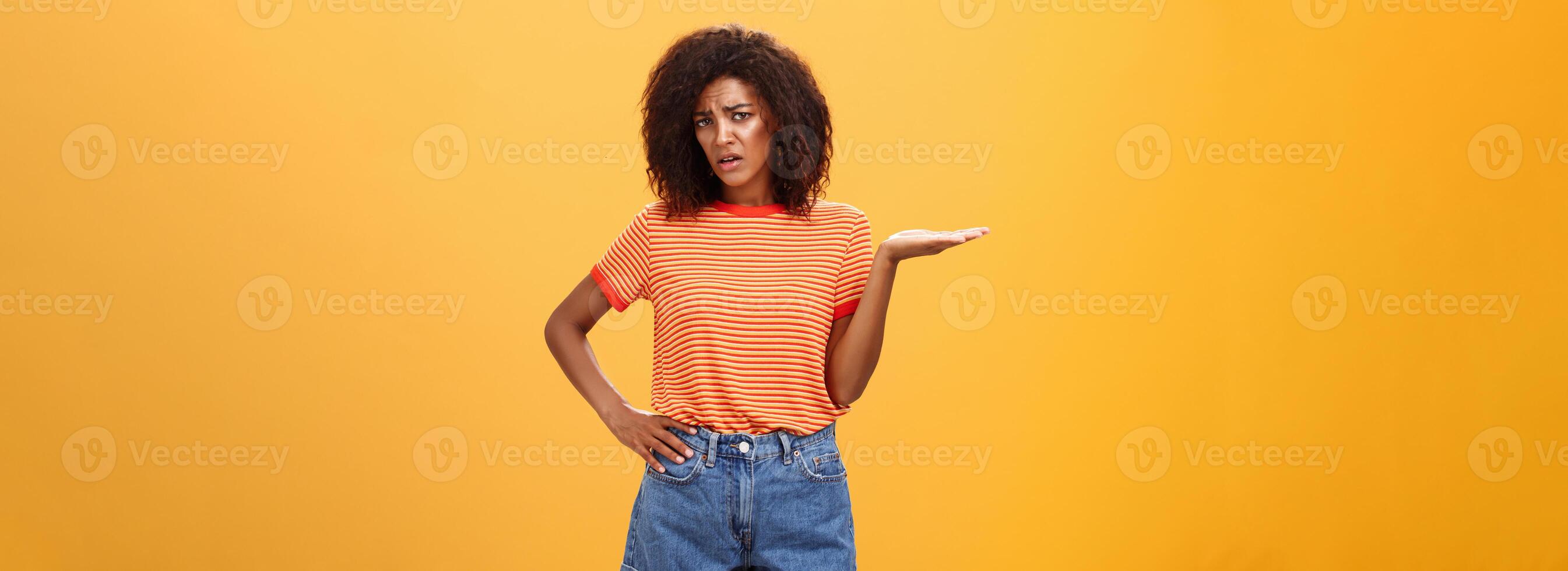 So what. Moody unimpressed stylish dark-skinned girlfriend with curly hairstyle holding hand on waist frowning and raising palm in your point gesture being confused and questioned over orange wall photo