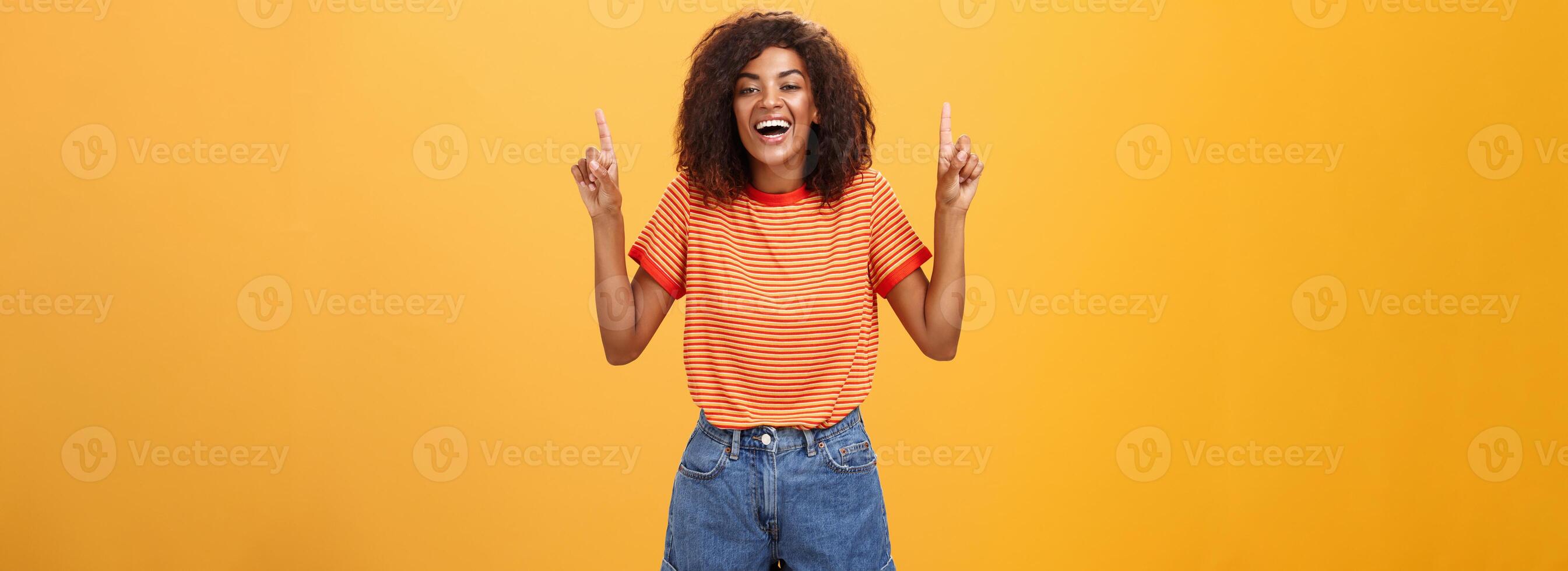 Woman feeling amused and entertained. Portrait of happy carefree stylish African-American girl with afro hairstyle laughing out loud joyfully pointing up with raised arms over orange wall photo