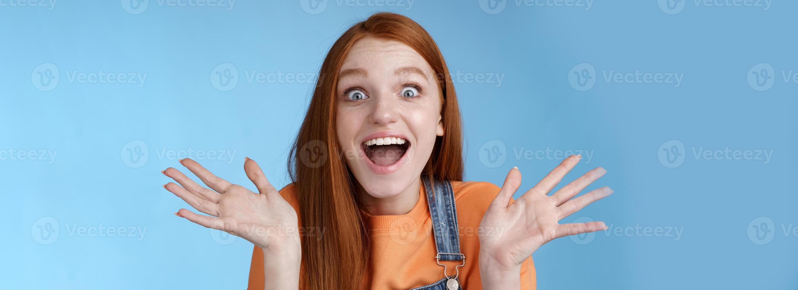Amused speechless surprised astonished redhead girl hear exciting positive news smiling gladly wide eyes thrilled raised hand triumphing win lottery celebrating victory success, blue background photo