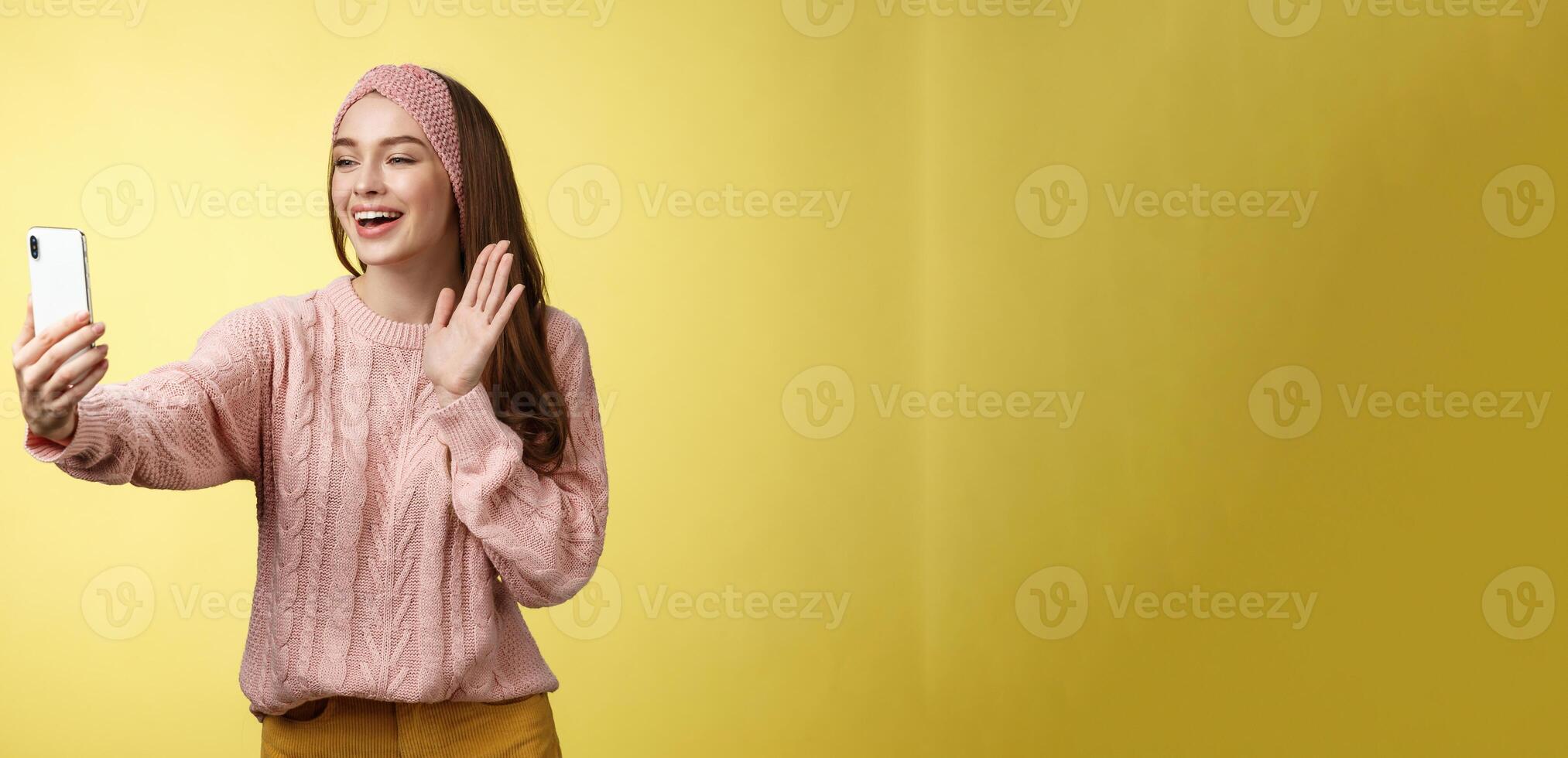 Charming outgoing girlfriend having online video chat, holding smartphone waving at phone camera saying hello talking via videocall smiling friendly during pleasant conversation, recording greeting photo