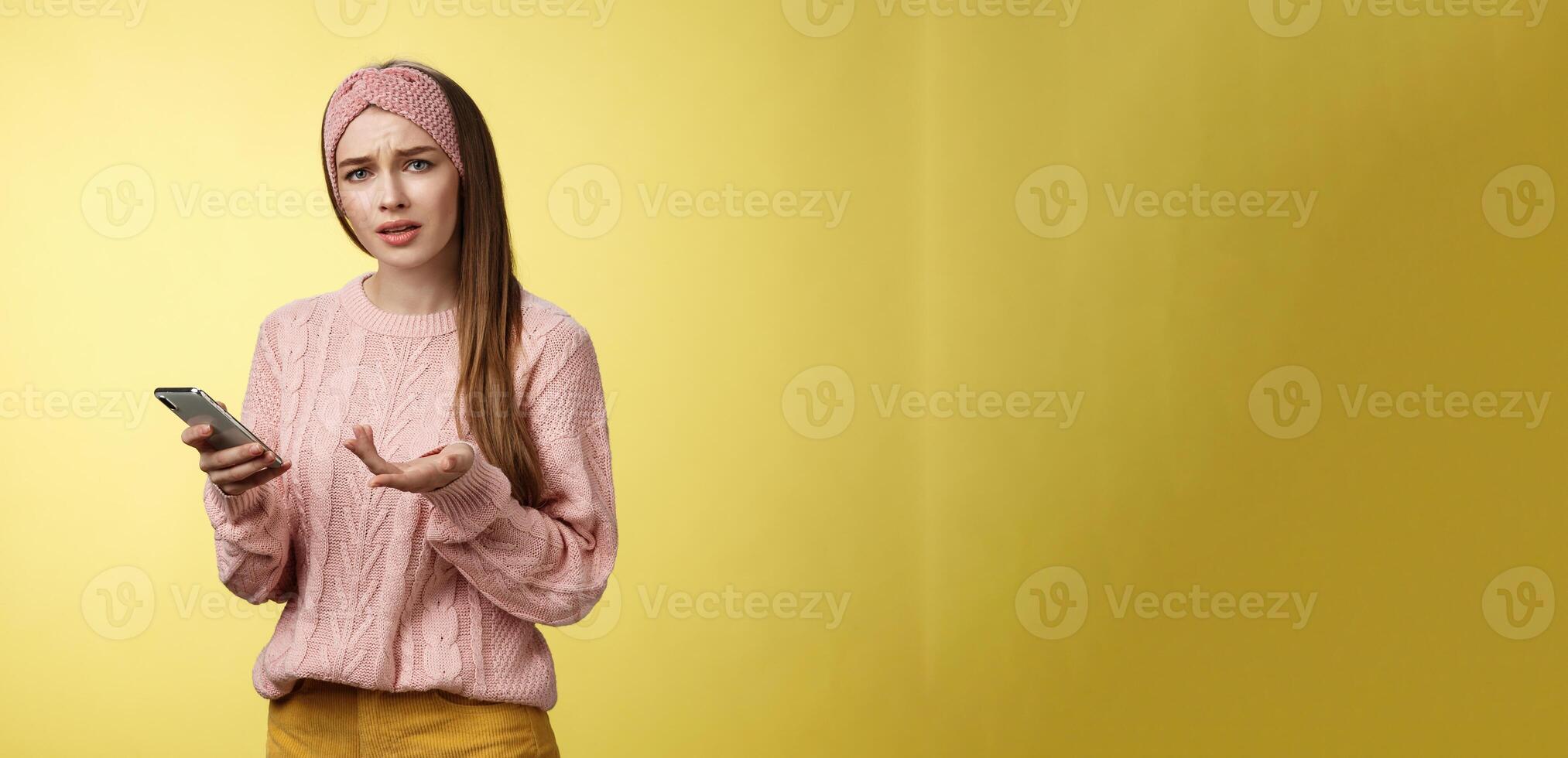 Puzzled confused upset questioned young cute girl in headband knitted sweater frowning clueless raising hand in dismay holding smartphone, cannot understand what happened cellphone over yellow wall photo