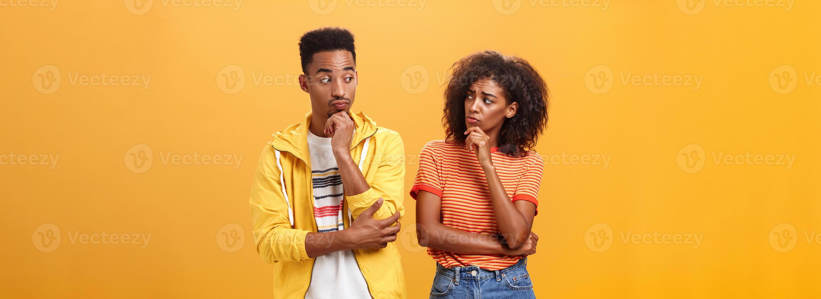 Silly confused girl standing gloomy and worried pursing lips and frowning holding hand on chin looking at boyfriend confused while guy glancing at girlfriend with curious expression over orange wall photo