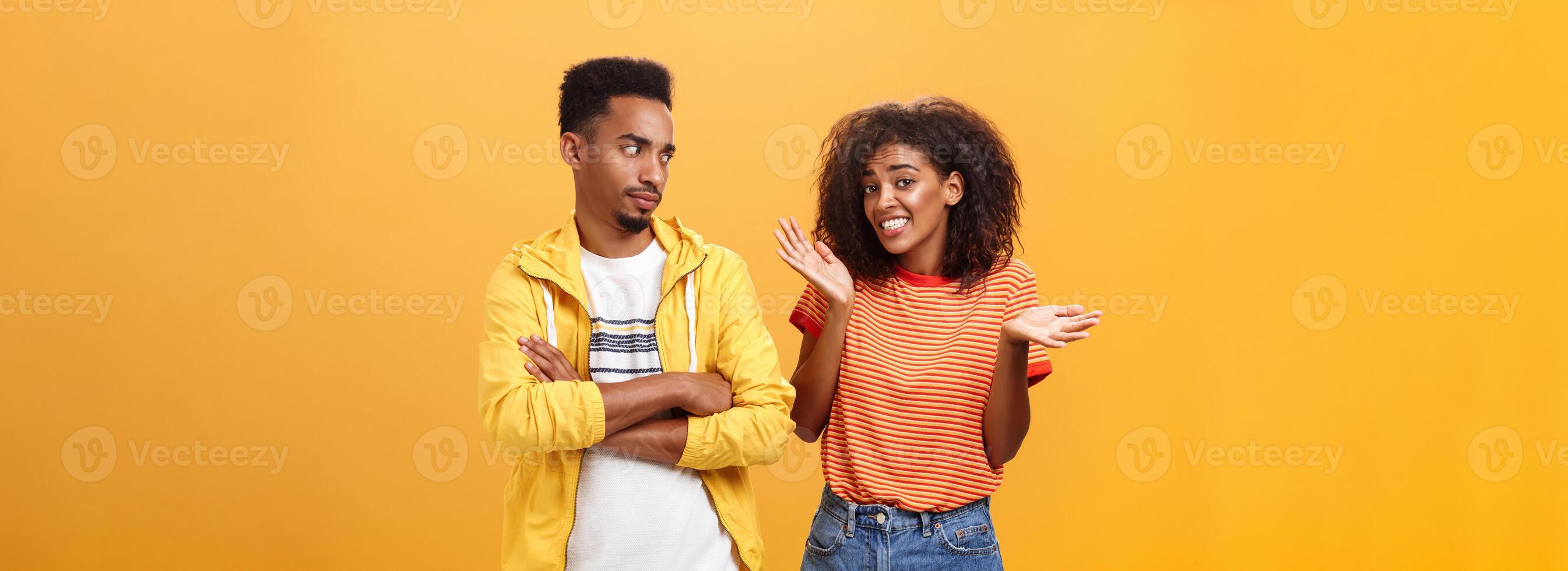Guy thinks his friend weirdo making dumb thinks looking at cute girl with suspicious look crossing arms on chest raising eyebrow questioned while girlfriend saying sorry shrugging over orange wall photo