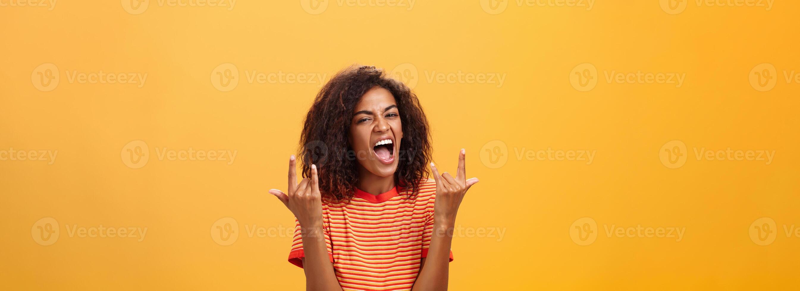Waist-up shot of amazed happy stylish african american woman feeling awesome rocking on party yelling from joy and satisfaction showing rock n roll gesture posing over orange background photo