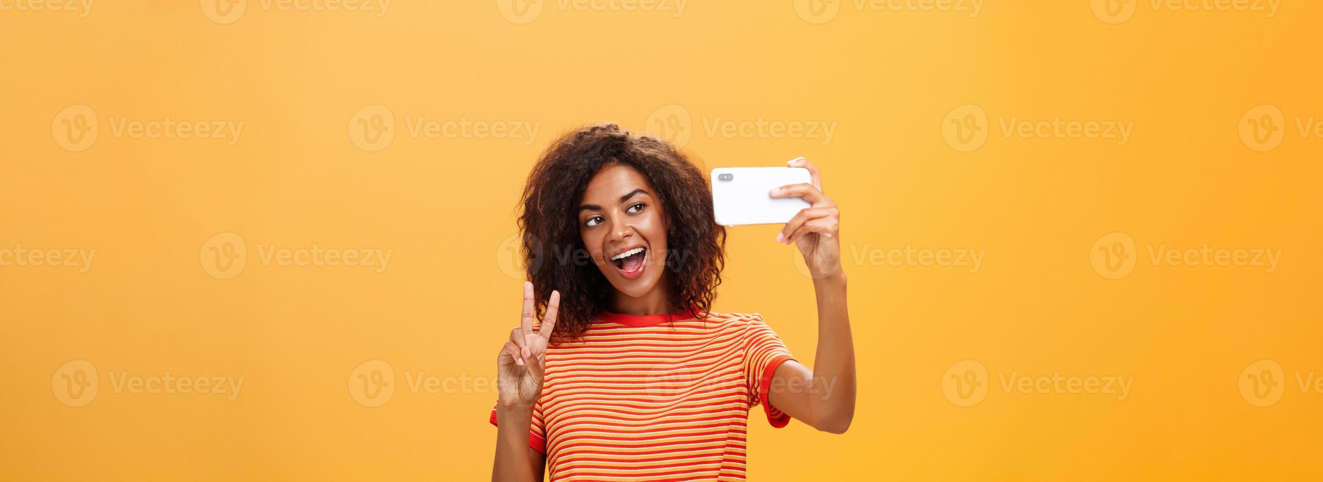 Smile to camera. Attractive and stylish self-assured dark-skinned female model with curly hairstyle showing peace gesture while taking selfie holding smartphone near face and smiling at gadget screen photo