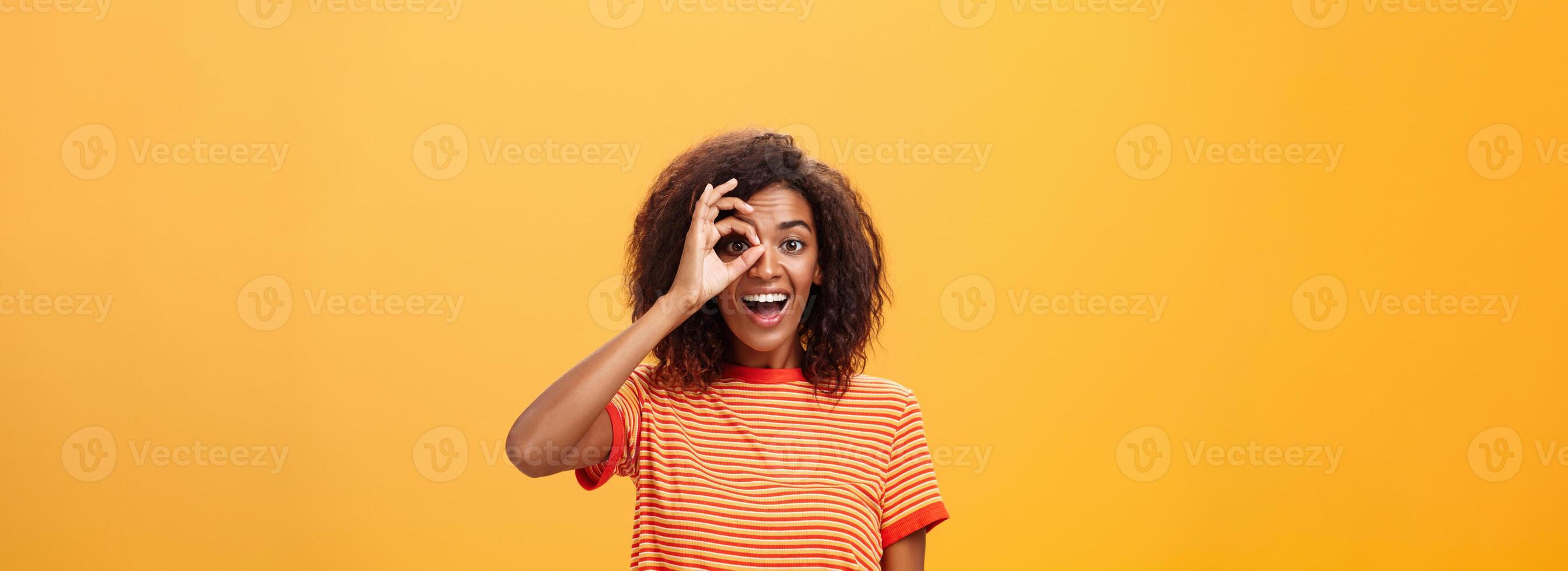 Creative and playful dreamy dark-skinned adult girl with curly hairstyle in striped t-shirt showing circle over eye or okay gesture smiling broadly ready take part in adventures near orange wall photo