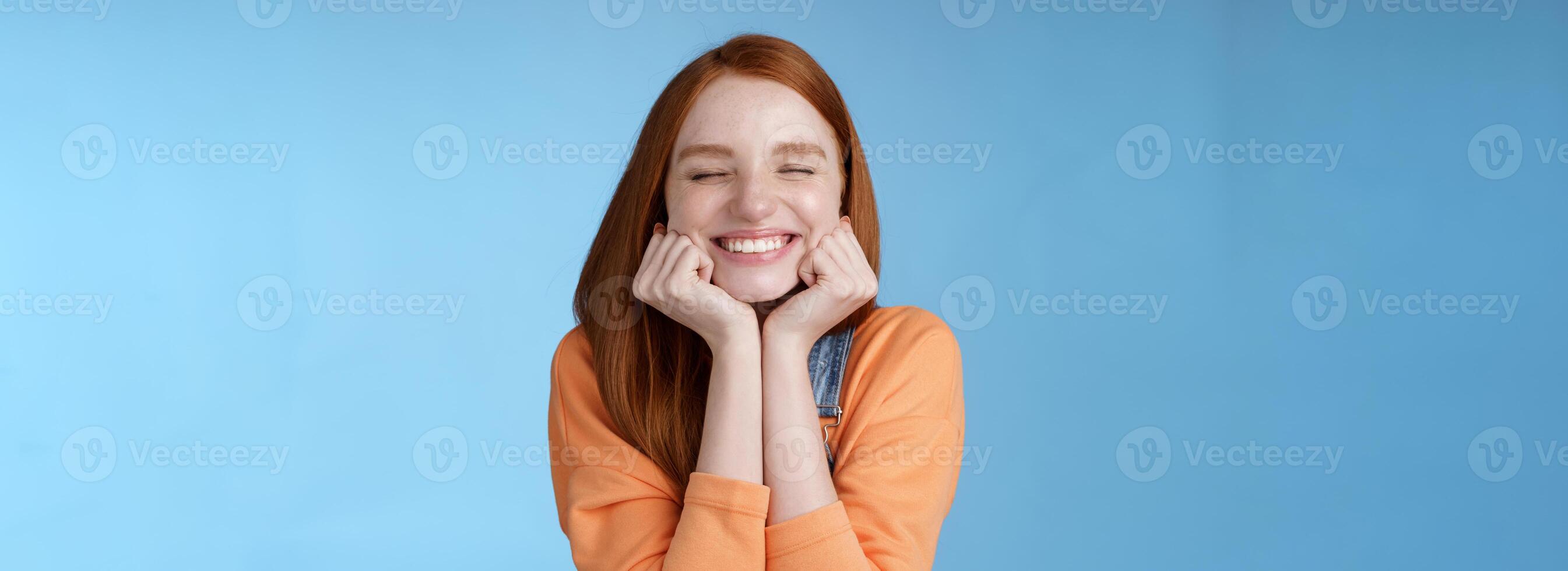 Silly cute happy redhead girlfriend smiling broadly close eyes dreamy squeez cheeks delighted asked date guy likes standing blue background rejoicing have fantastic lucky day triumphing photo