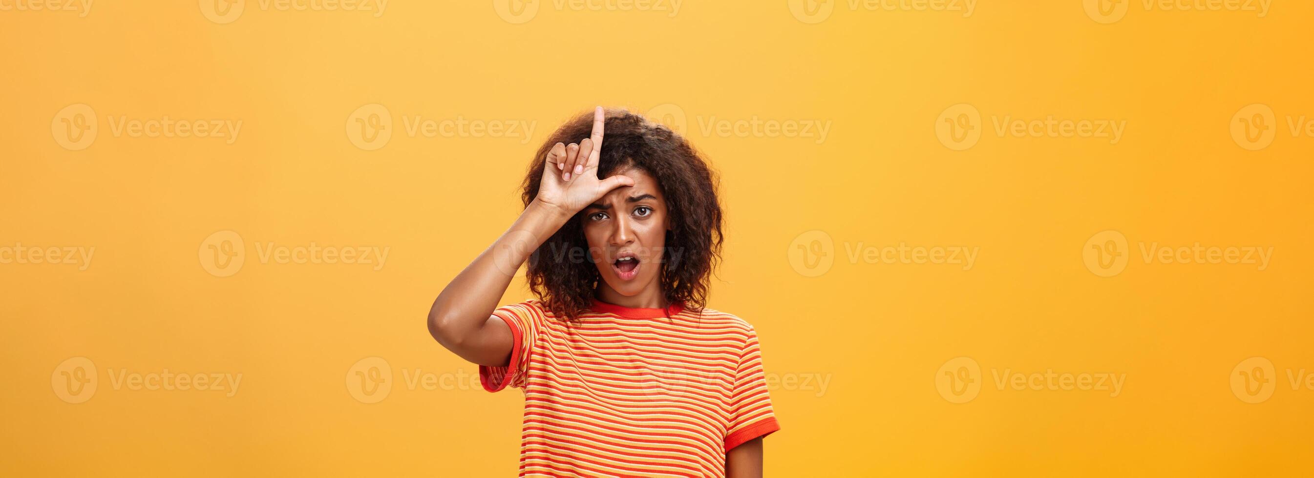 Girl thinks she loser. Portrait of gloomy bothered and displeased african american woman with afro hairstyle showing l word over forehead complaining feeling gloomy and unhappy over orange background photo