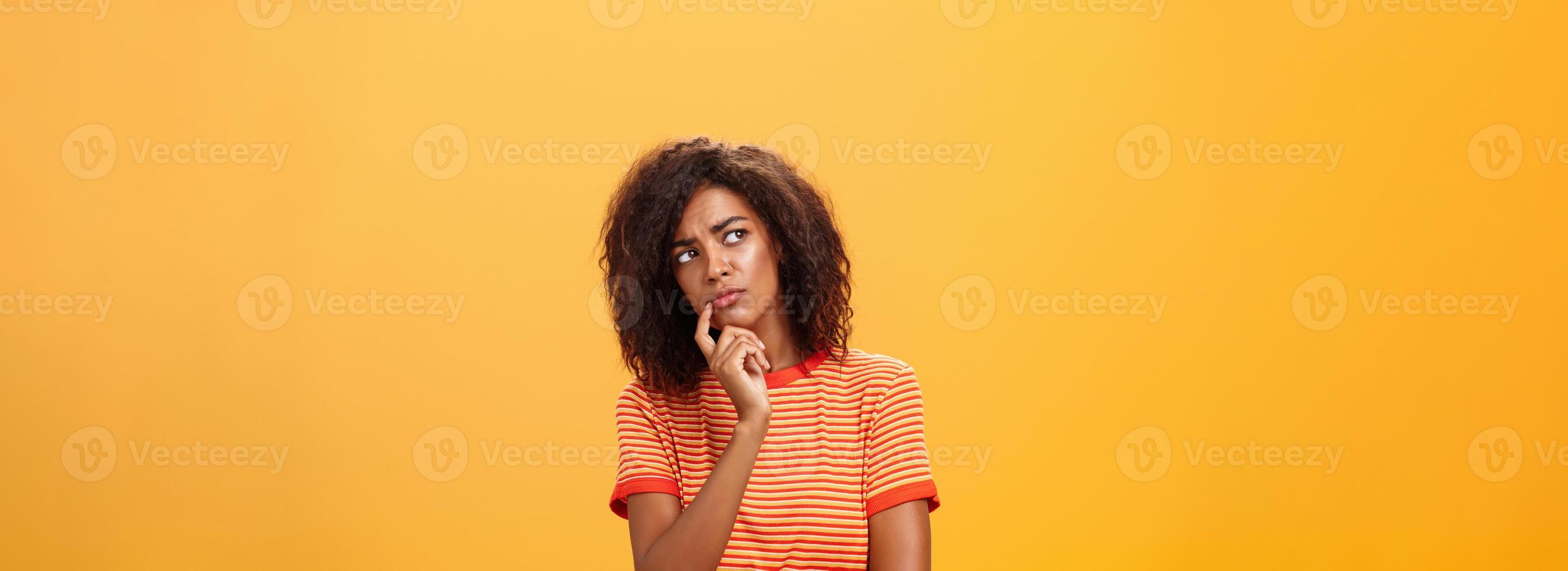 Woman considering to take part in event. Smart focused and creative good-looking african girl with curly hair holding finger on lip gazing concerned left, thinking making decision over orange wall photo