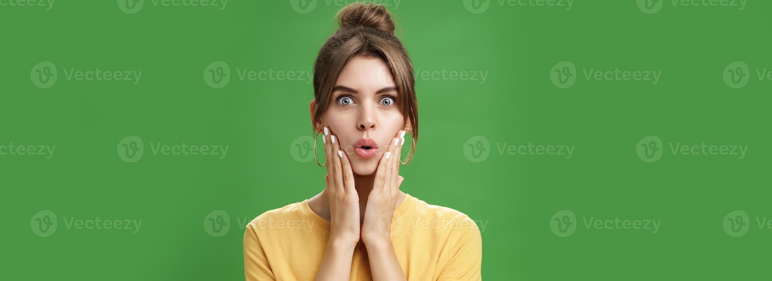 Portrait of impressed and shocked cute feminine woman in yellow t-shirt folding lips from excitement and interest touching cheeks surprised reacting to amazing rumor posing over green background photo