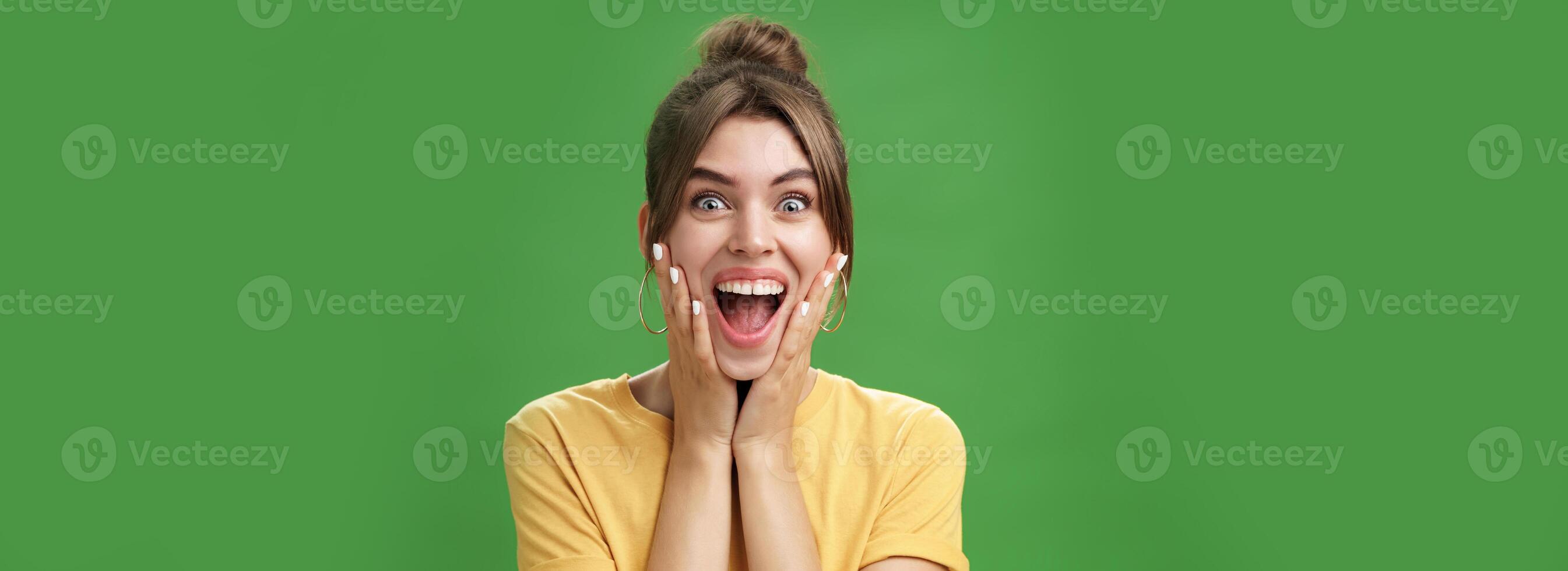 Portrait of happy delighted and surprised young feminine girl in yellow t-shirt pressing hands to cheeks from amazement and joy smiling broadly reacting to astonishing news over green background photo