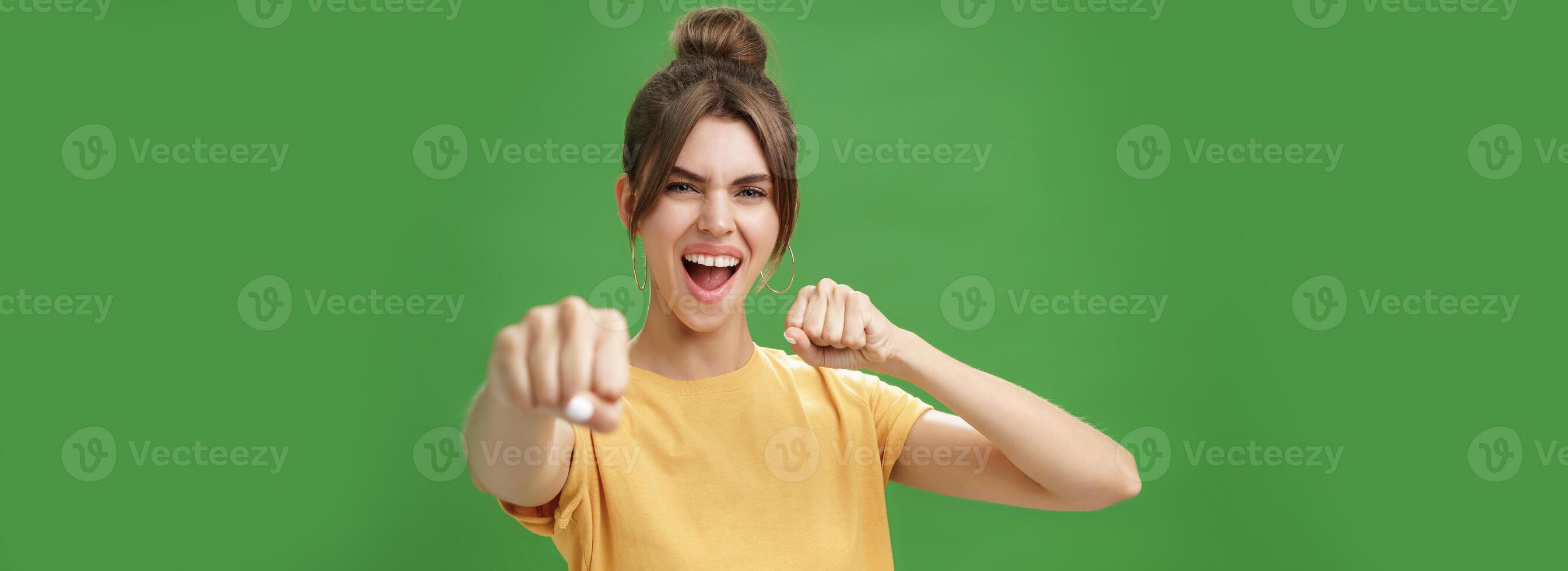 Cute female rebel in yellow t-shirt with gap teeth pulling fist towards camera as if showing fighting skills yelling daring and excited standing over green background smiling acting like boxer photo