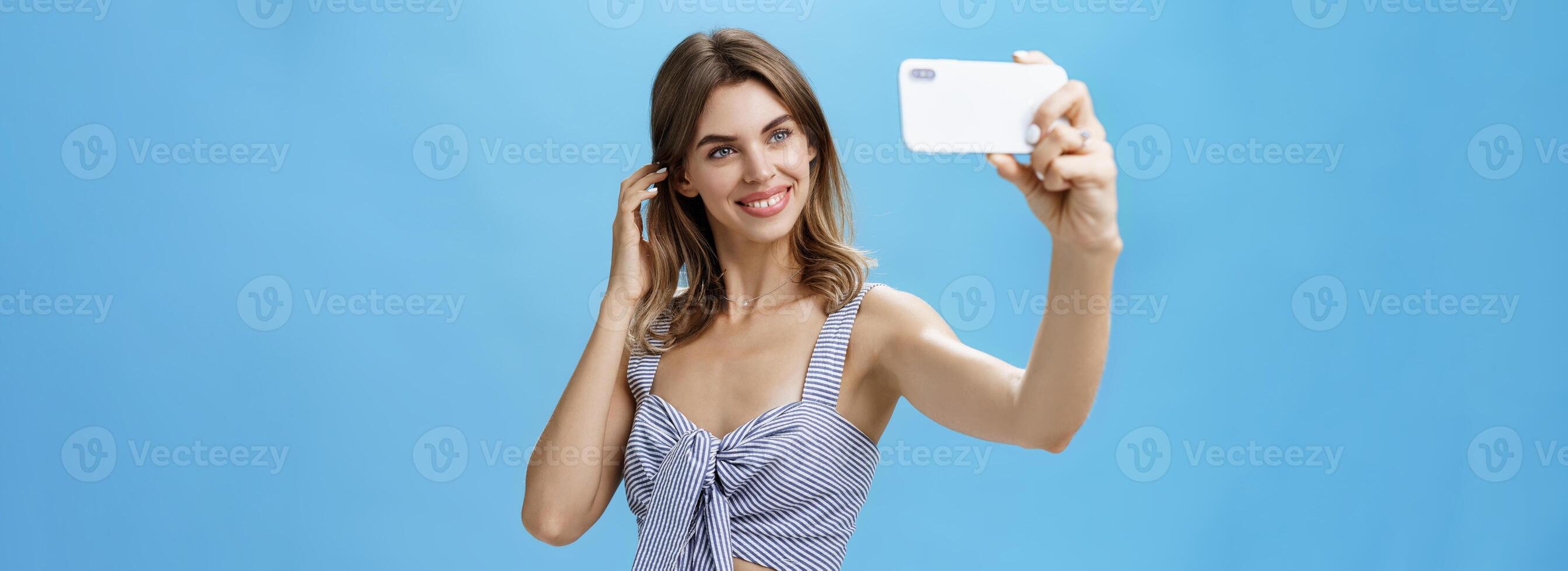 atractivo mujer con bueno autoestima en elegante pareo atuendo sacudiendo pelo detrás oído sonriente alegremente a teléfono inteligente pantalla, tomando selfie a enviar en Internet y ganancia seguidores terminado azul pared foto