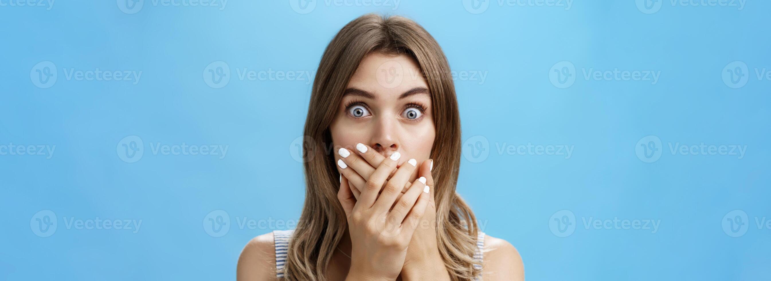 Close-up shot of shocked speechless woman covering mouth with pressed palms popping eyes at camera from surprise and worry, gasping witnessing terrible scene, feeling scared and intense over blue wall photo