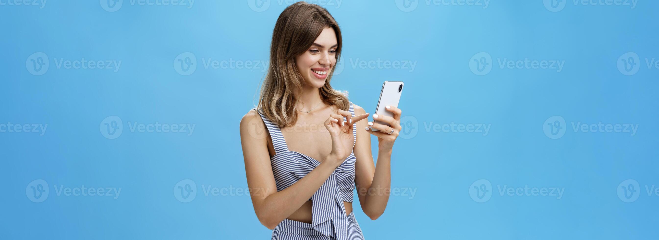 Portrait of stylish sociable good-looking female entrepreneur making appointment via smartphone holding cellphone typing message gazing at device screen with pleased carefree smile over blue wall photo