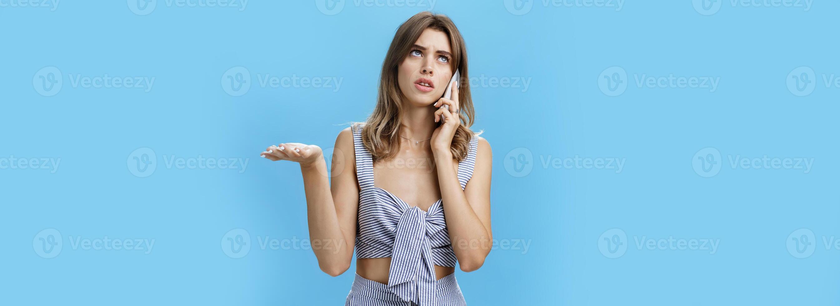 Waist-up shot of perplexed and confused cute girlfriend in matching outfit making shoulder shrug and raising palm in clueless gesture looking up while talking on phone cannot understand what answer photo