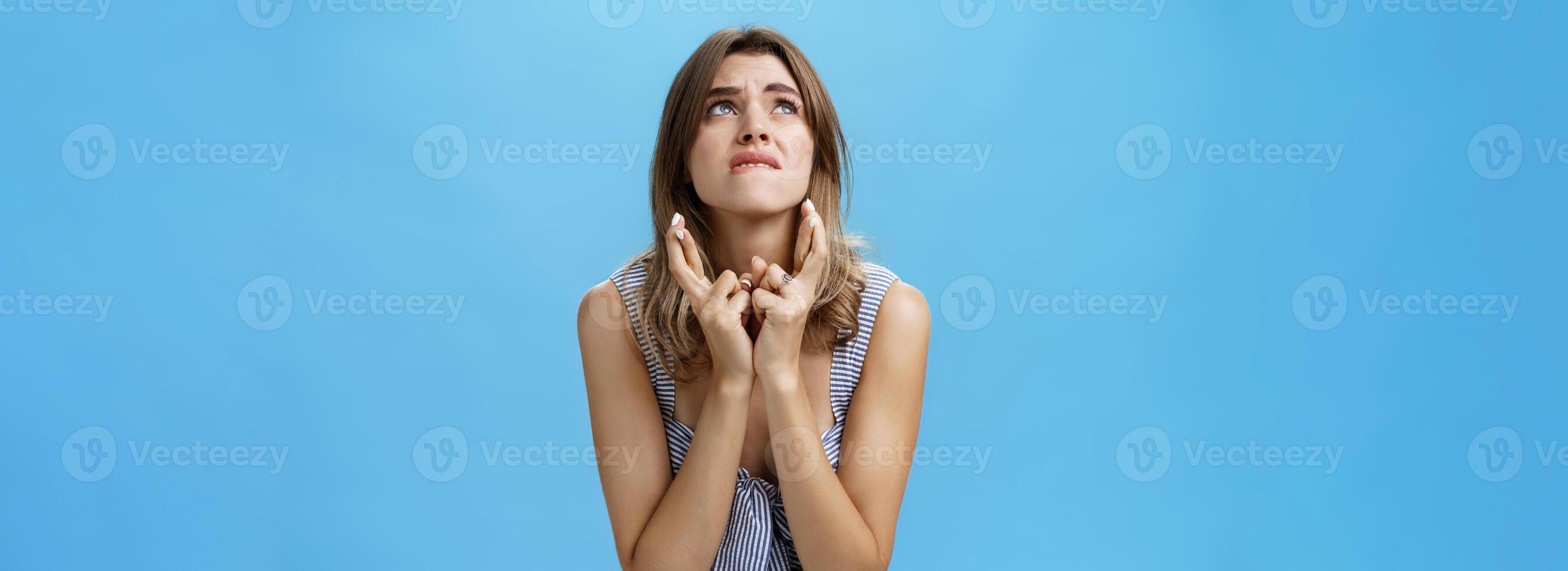 Waist-up shot of intense cute young woman crossing fingers biting lower lip and frowning stooping shoulders looking up concerned and hopeful waiting for miracle praying for good luck over blue wall photo