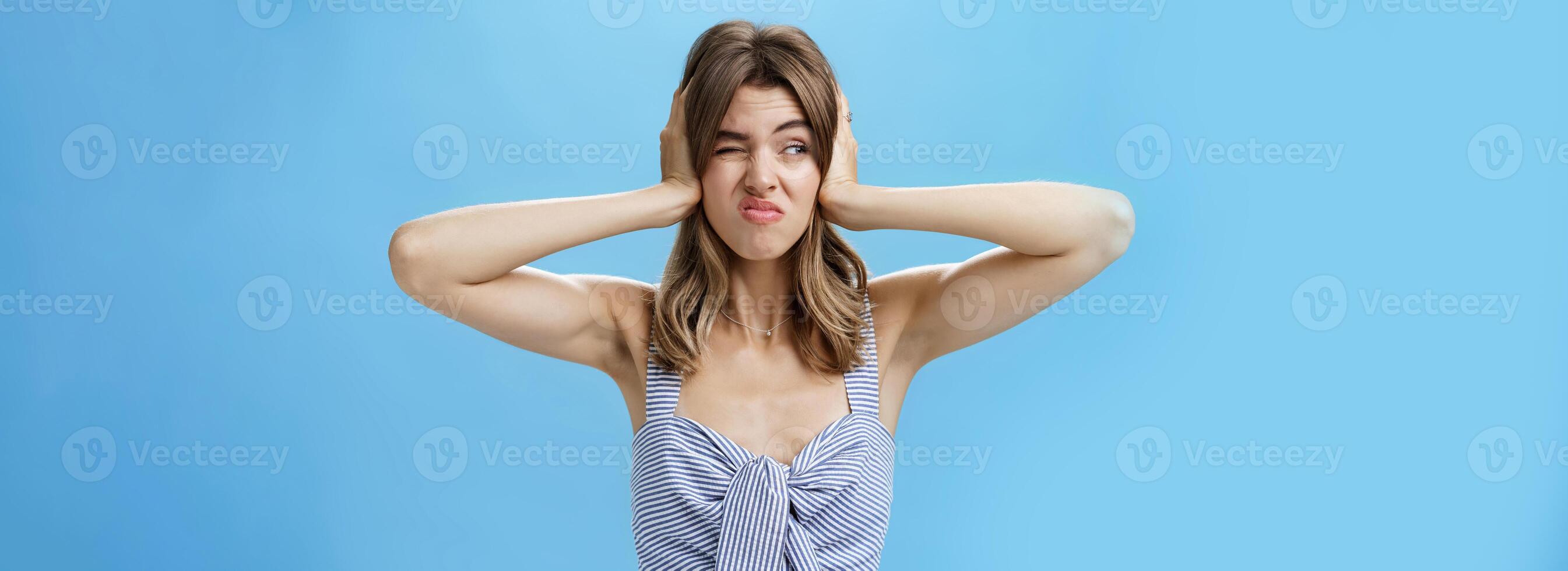 Girl feeling irritated by loud awful noise pressing hands to ears not hear annoying sound of drill smirking and frowning, squinting looking left displeased and disturbed, posing over blue background photo