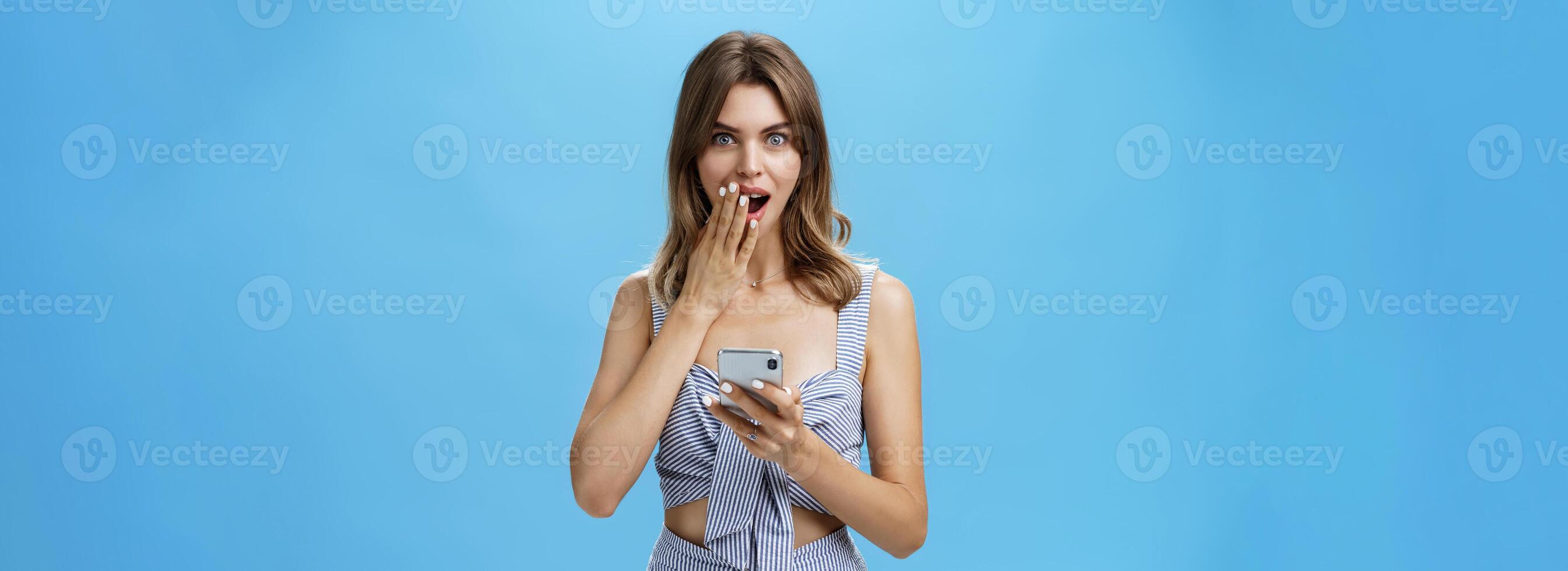 Waist-up shot of surprised and excited attractive woman holding smartphone covering opened mouth with palm after reading shocking rumor in social-network via cellphone against blue background photo