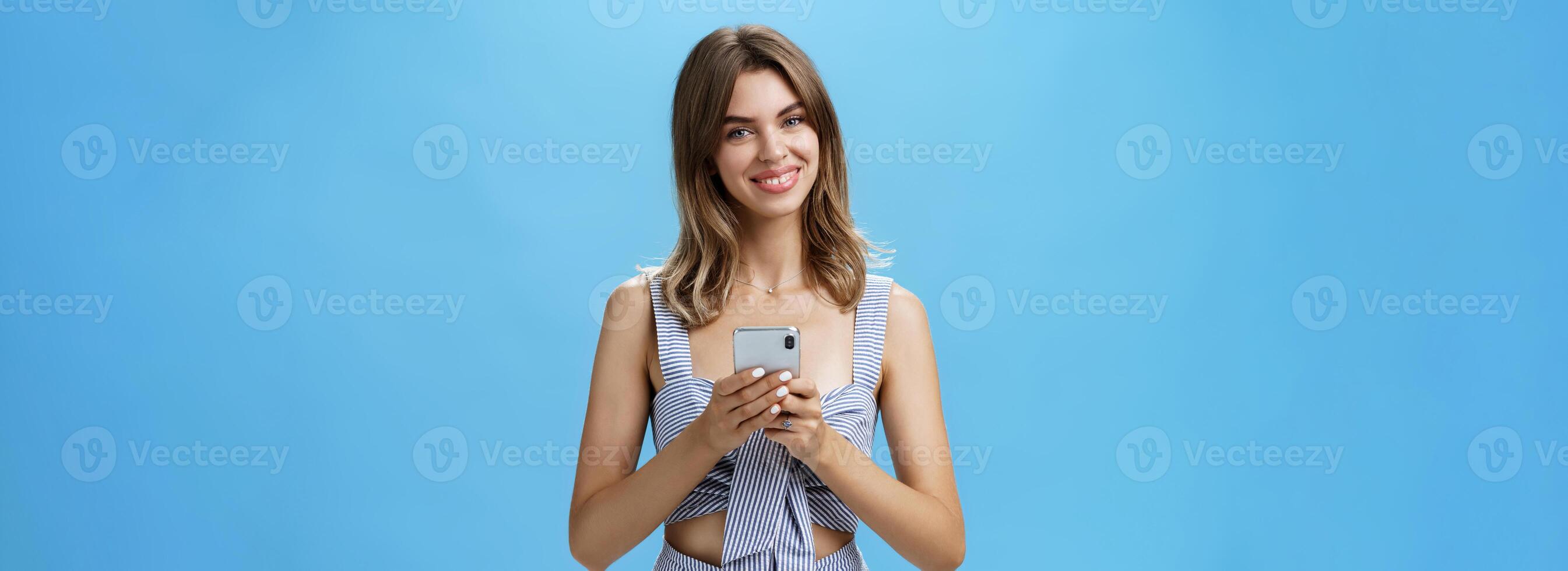 Friendly pleasant good-looking woman in matching outfit holding smartphone over chest tilting head smiling broadly showing cute gapped teeth being delighted after reading heartwarming message photo