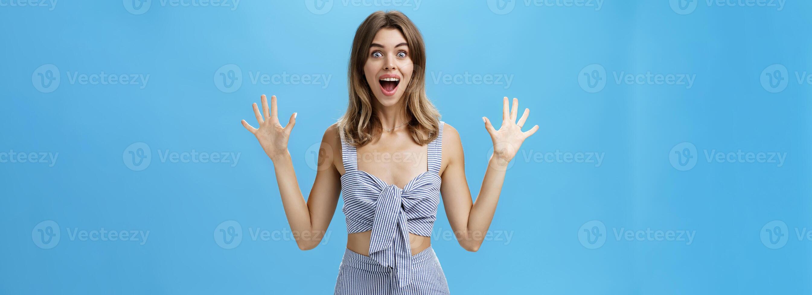 Cute woman with gapped teeth feeling exciting from surprise and anticipation smiling broadly raising palms from thrill and amazement popping eyes at camera posing over blue background photo