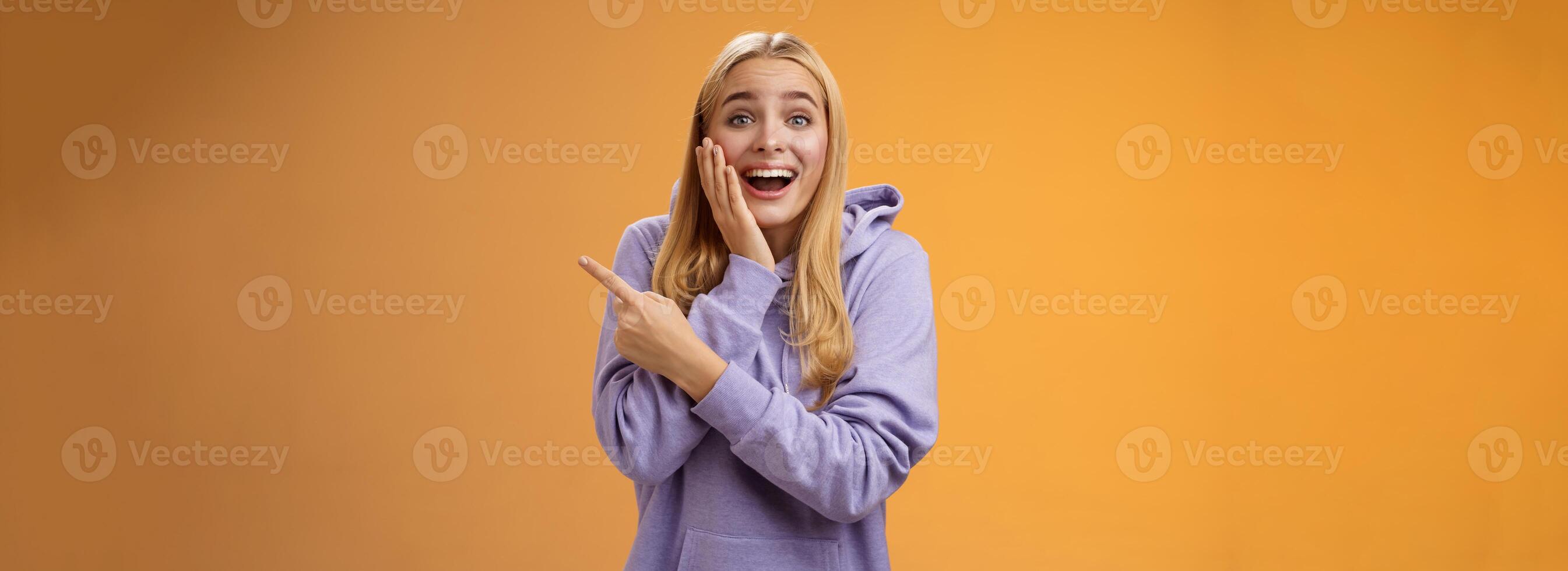 Amused excited speechless happy young blond woman 25s in hoodie cannot believe luck happiness pointing right amazed smiling broadly touch cheek surprised receive awesome unexpected gift photo