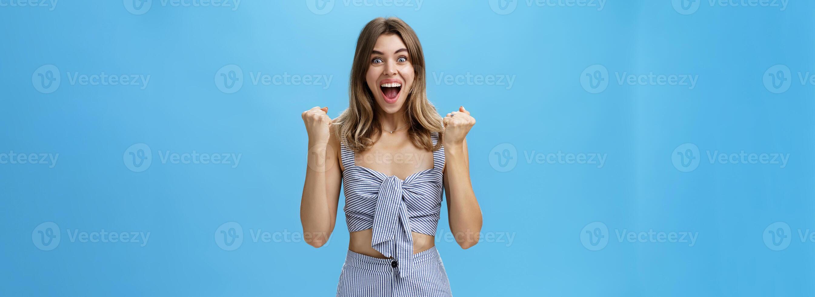 Happy surprised and triumphing young nice girl with wavy natural hairstyle in matching striped outfit raising clenched fists in cheer and joy celebrating success or fortune smiling broadly at camera photo