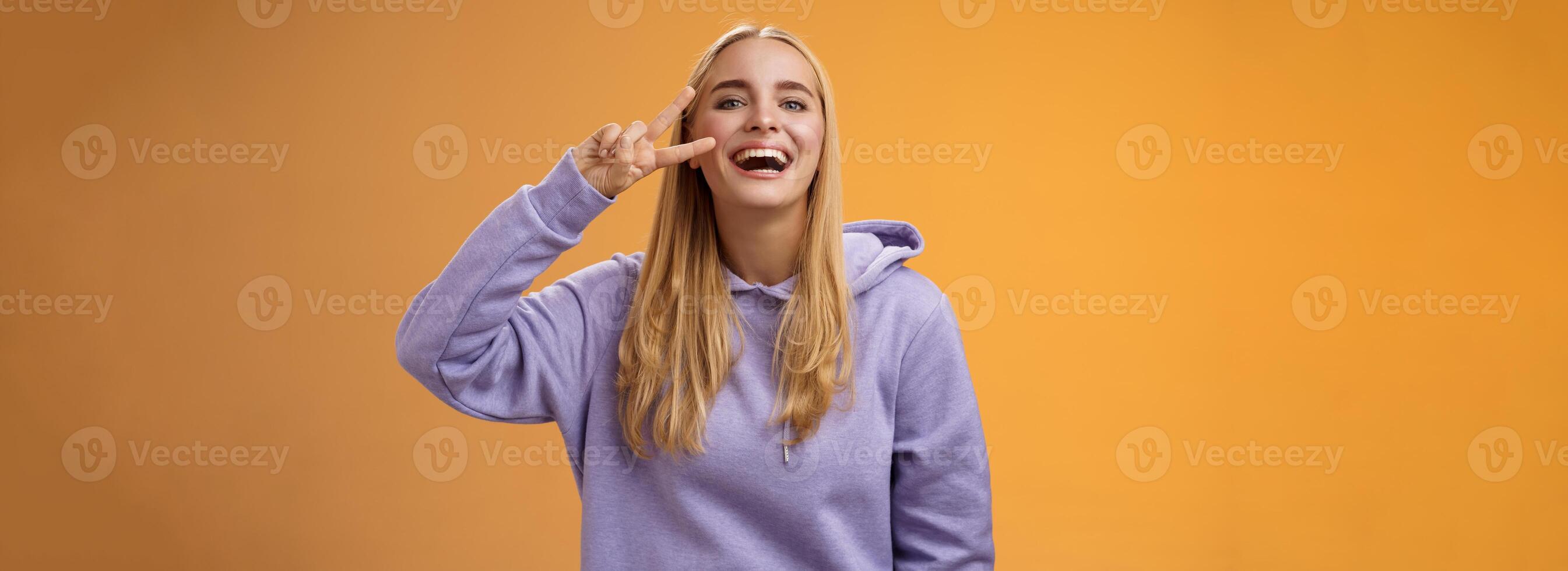 Attractive friendly carefree millennial blond girl in purple hoodie having fun friends laughing joyfully show peace victory hippie sign adore perfect chilly spring weather, orange background photo