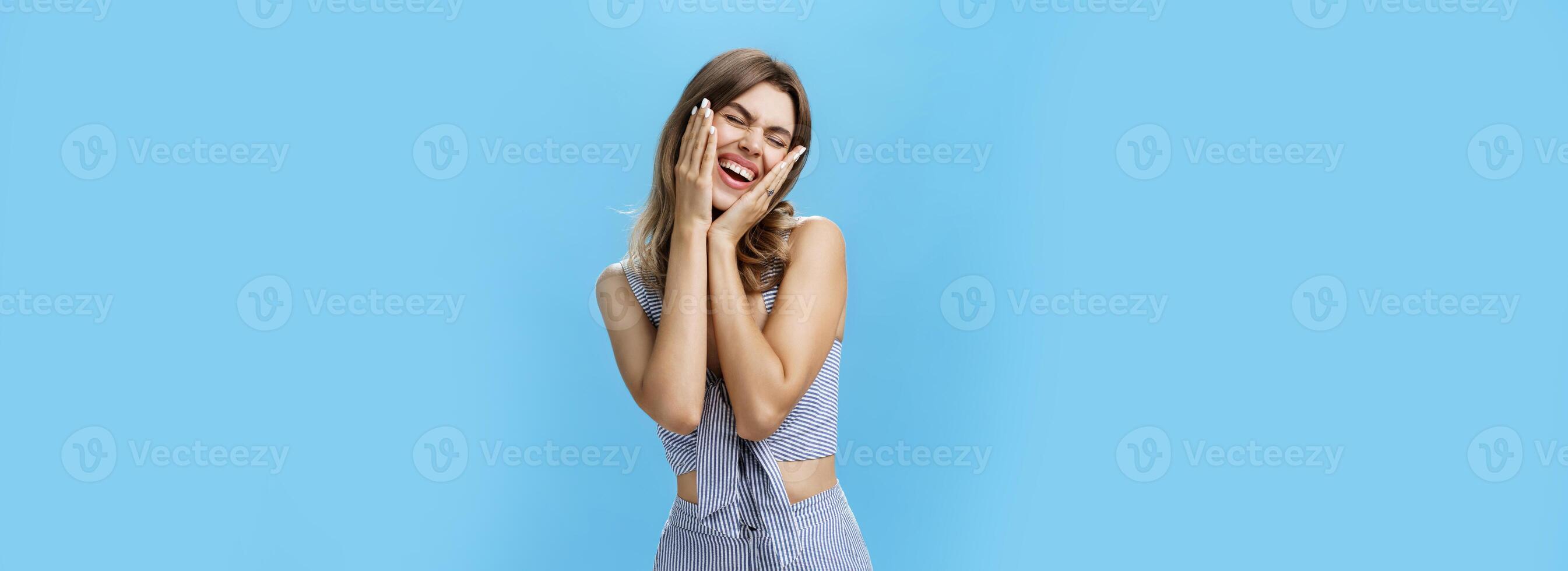 Studio shot of attractive charismatic female with natural beauty and gapped teeth closing eyes in joy and excitement pressed hands to cheeks from amazement and happiness posing against blue background photo