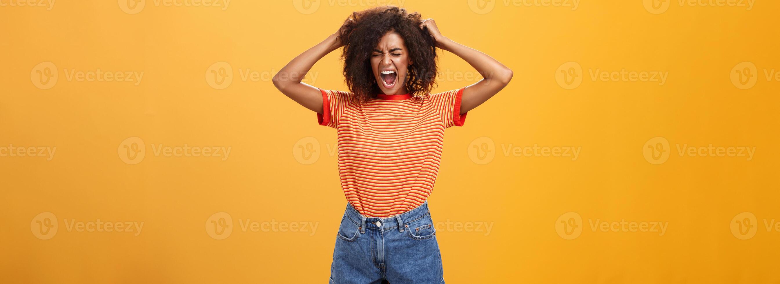 I hate life being cruel. Depressed african woman in rage and fury pulling hair out of head yelling and screaming with closed eyes losing temper having bad day hating everything over orange wall photo