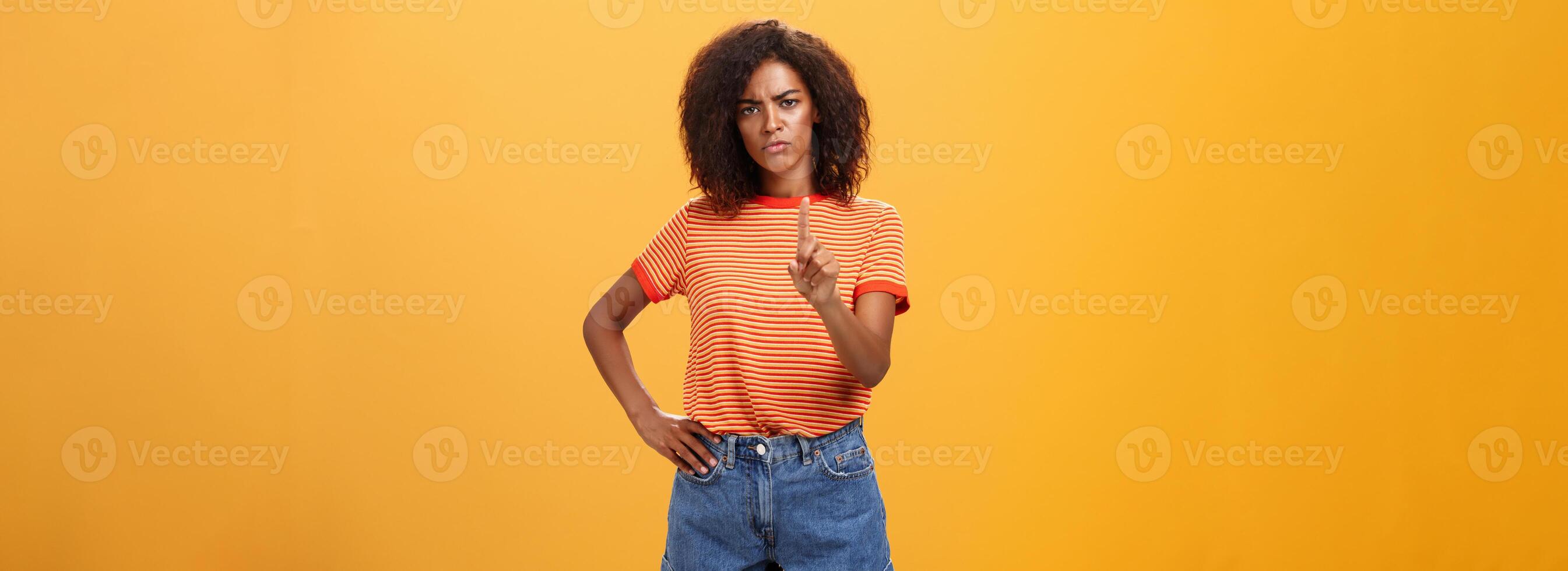 You better not. Dissatisfied bossy serious-looking african american curly-haired female head manager in t-shirt frowning shaking index finger in forbid or stop gesture scolding dog over orange wall photo