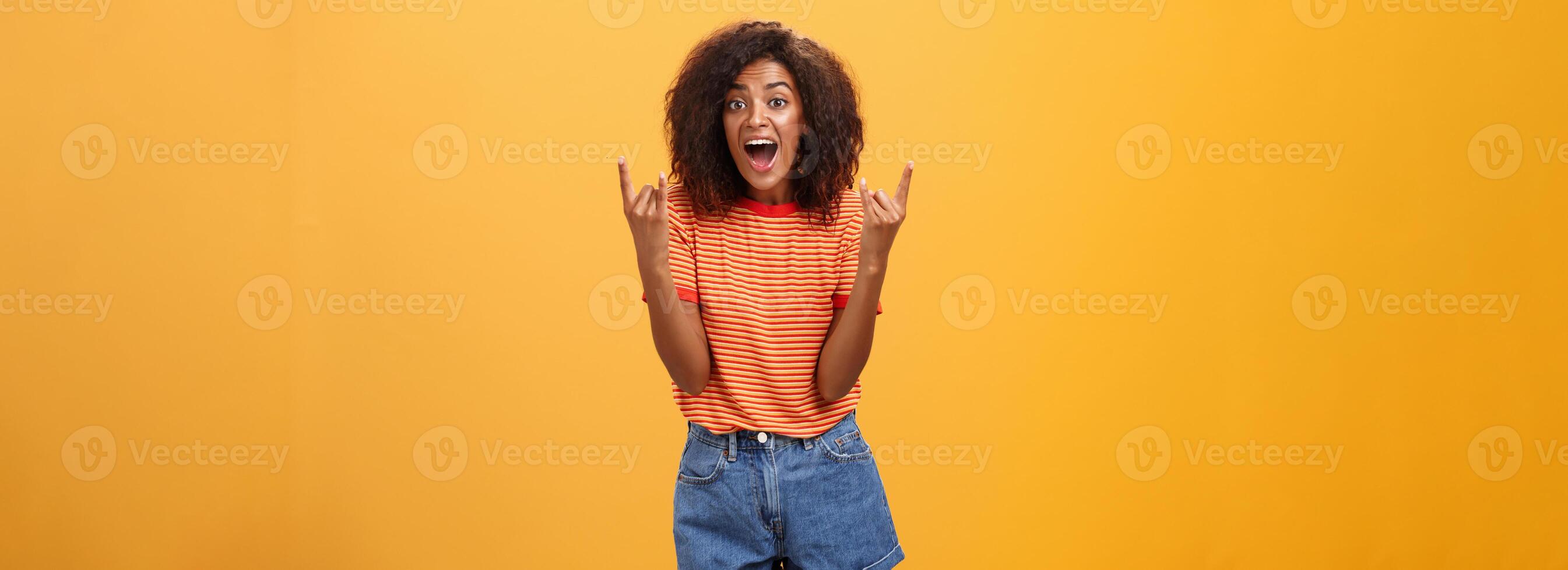 Stylish african american woman being delighted and excited before cool concert showing rock n roll gesture with both hands screaming from satisfaction and happiness cheering over orange background photo