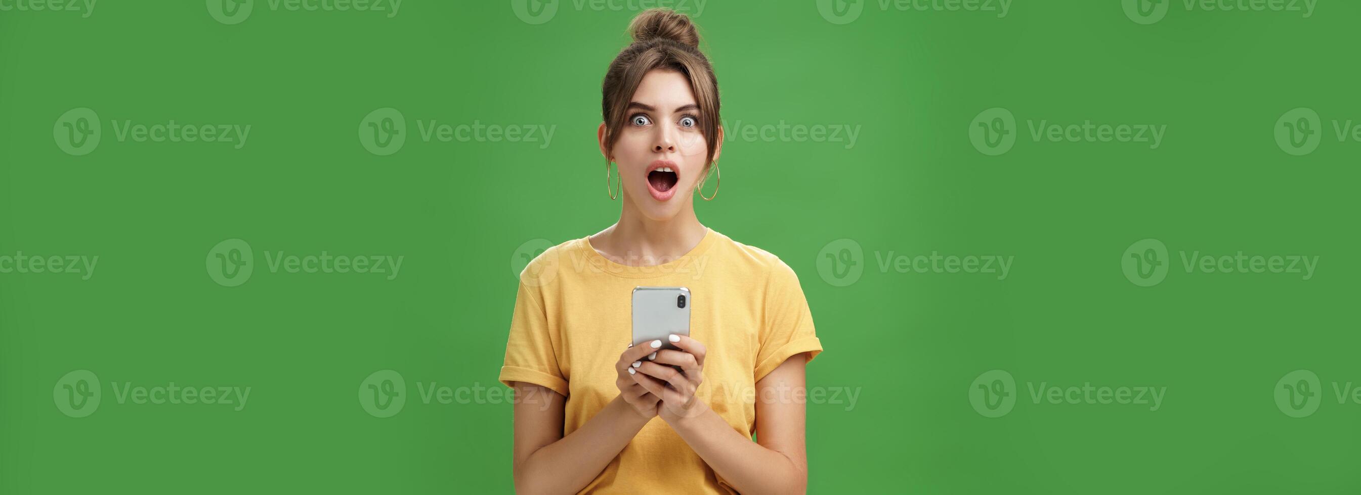 Portrait of shocked speechless and impressed beautiful white girl with combed hair in yellow t-shirt holding smartphone, dropping jaw from excitement reacting to cool app over green background photo