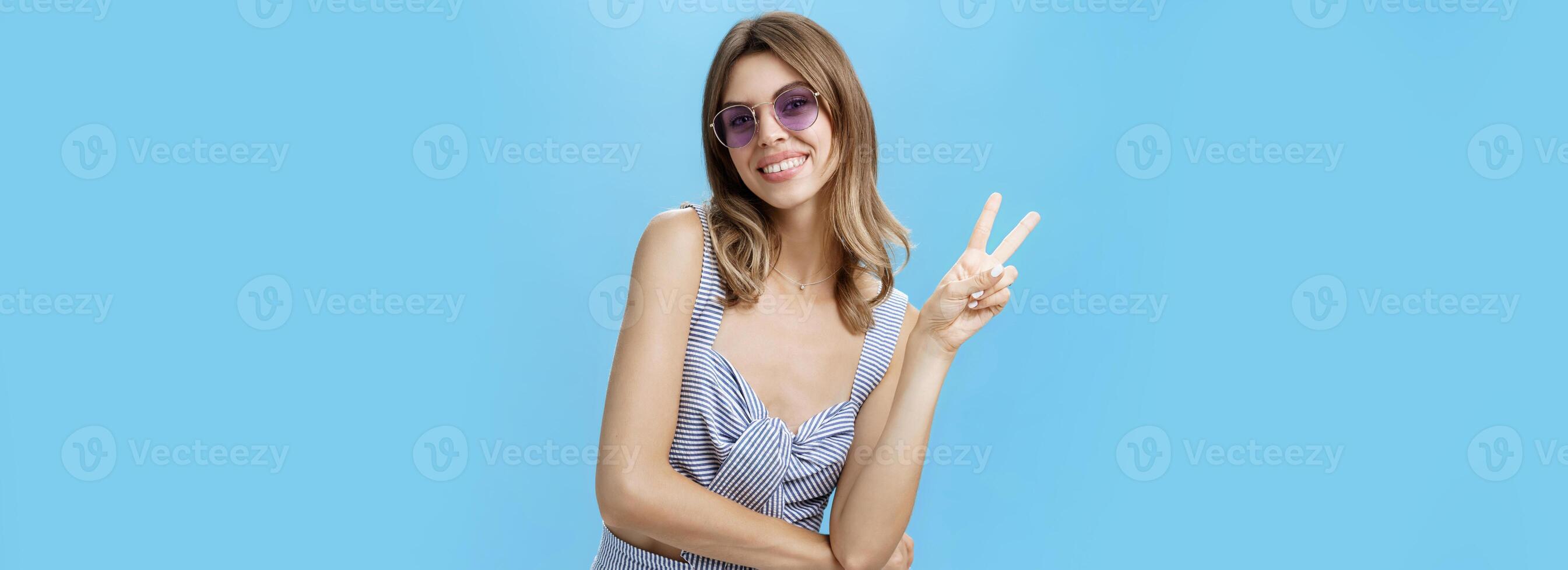 Charming friendly and outgoing young lady showing peace or victory gesture and smiling broadly at camera showing cute gapped teeth, tilting head and raising one shoulder posing flirty over blue wall photo