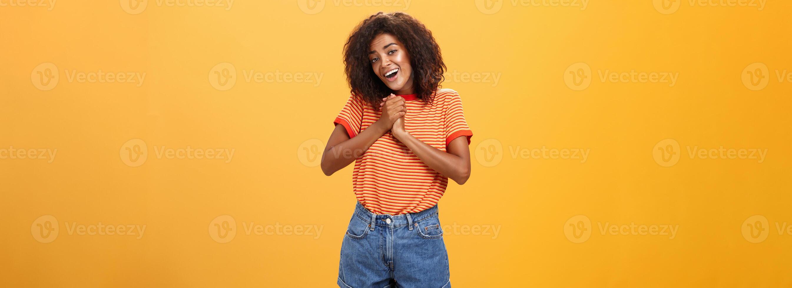 Girl sighing and gazing at cute thing clenching palms together near heart smiling with admiration and love being feminine and tender posing over orange background feeling affection photo